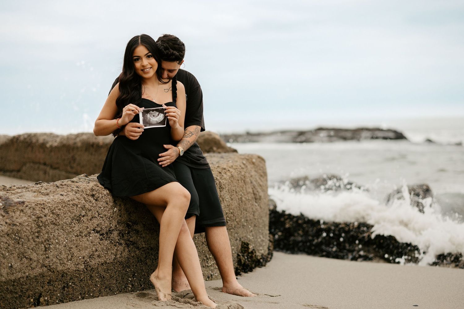 Romantic couple sitting on coastal rocks holding ultrasound photo at beach.