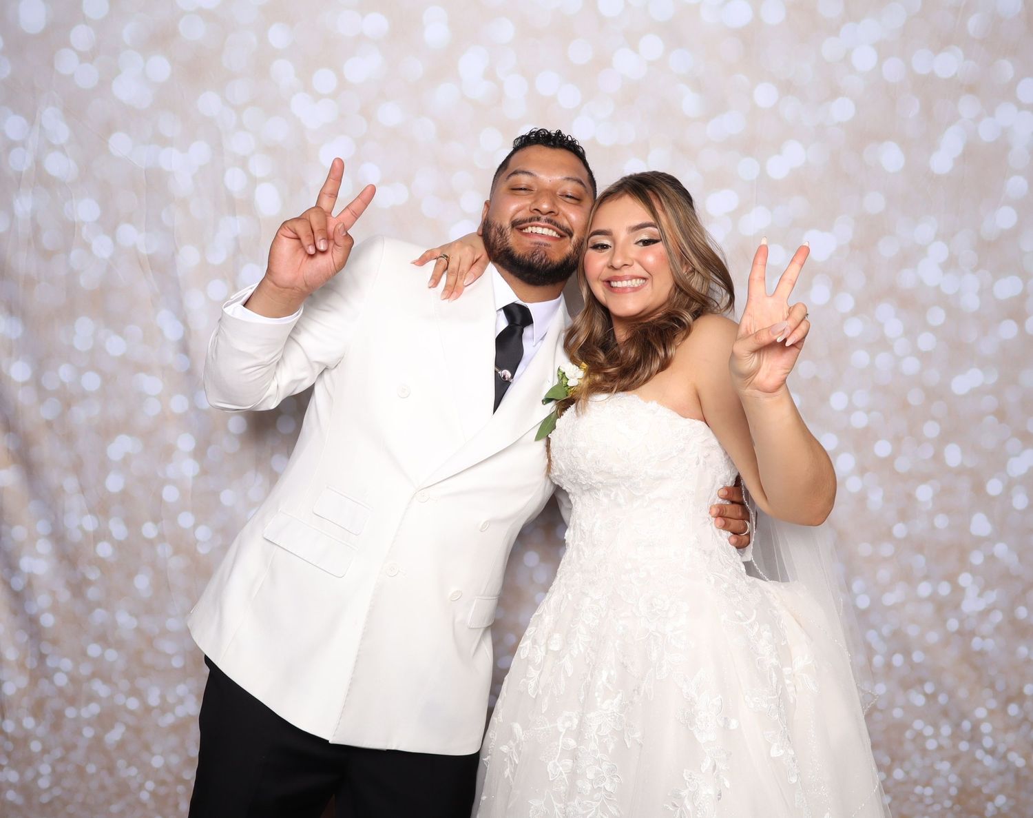Bride and groom in white wedding attire posing with peace signs in front of a bokeh backdrop.