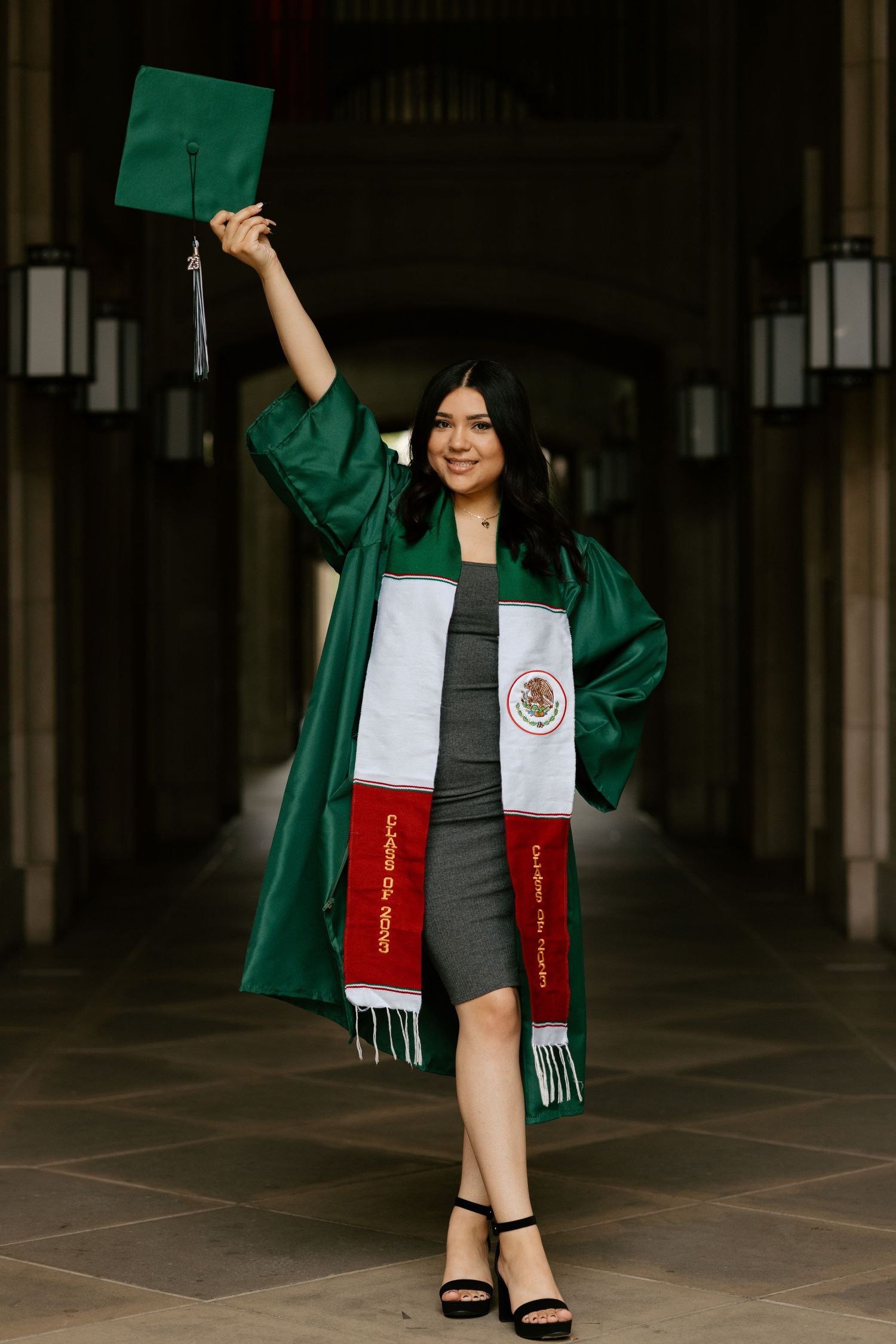 Graduate in emerald green gown holds cap aloft wearing Mexican flag colored stole.