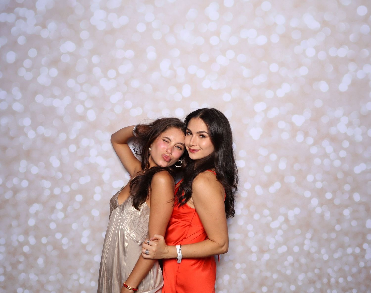 Two young women posing together in front of a bokeh light backdrop, one in gold and one in red dress.