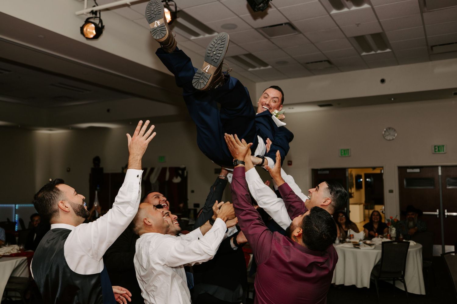 Large group of wedding guests dancing and celebrating with arms raised in a banquet hall.