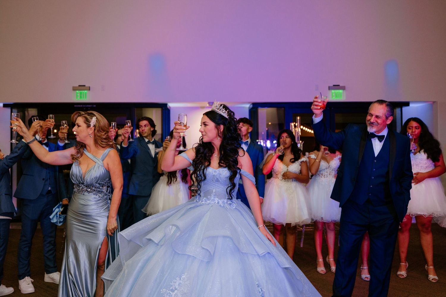 Festive ballroom celebration with elegant gowns and formal attire under purple lighting.