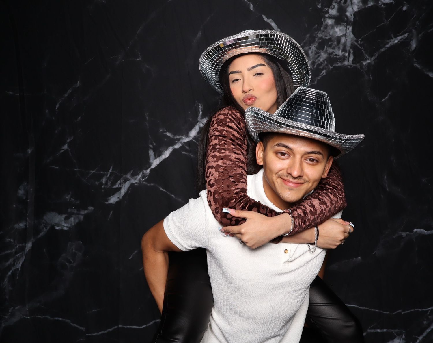 Two friends wearing sparkly cowboy hats pose playfully, one giving the other a piggyback ride against a marble backdrop.