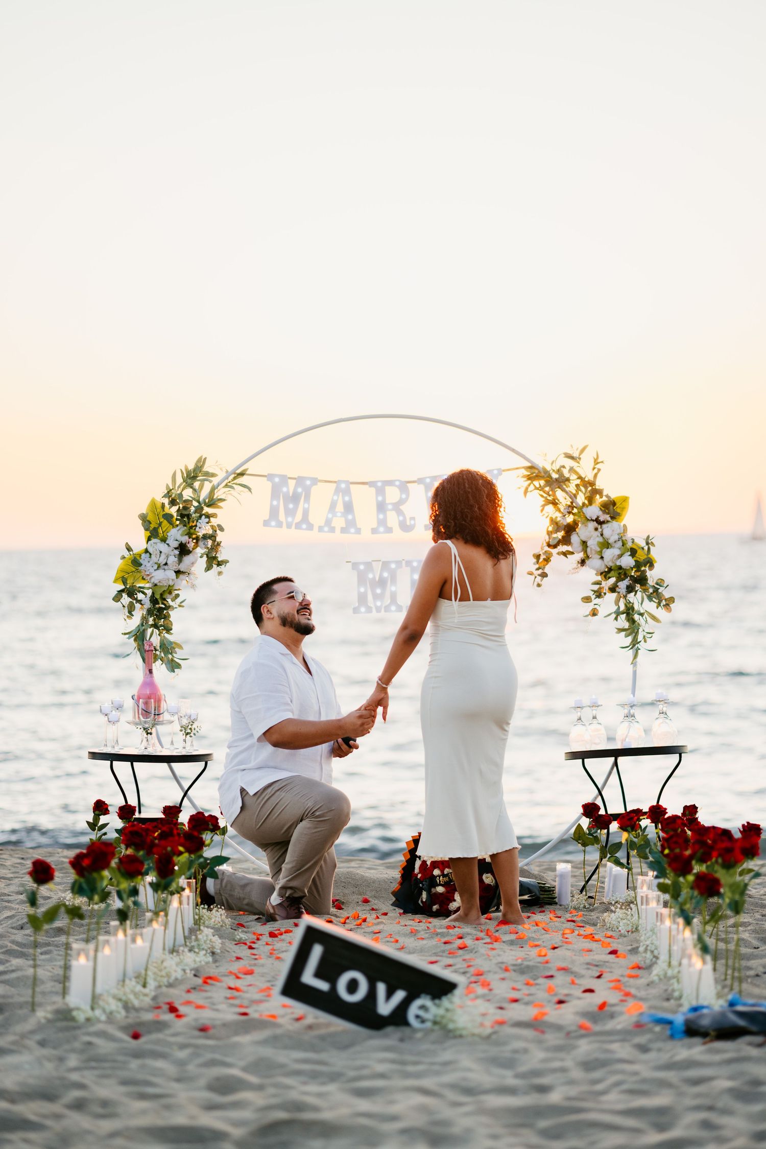 A romantic beach proposal setup with floral arch, rose petals, and a 'LOVE' sign during golden hour.