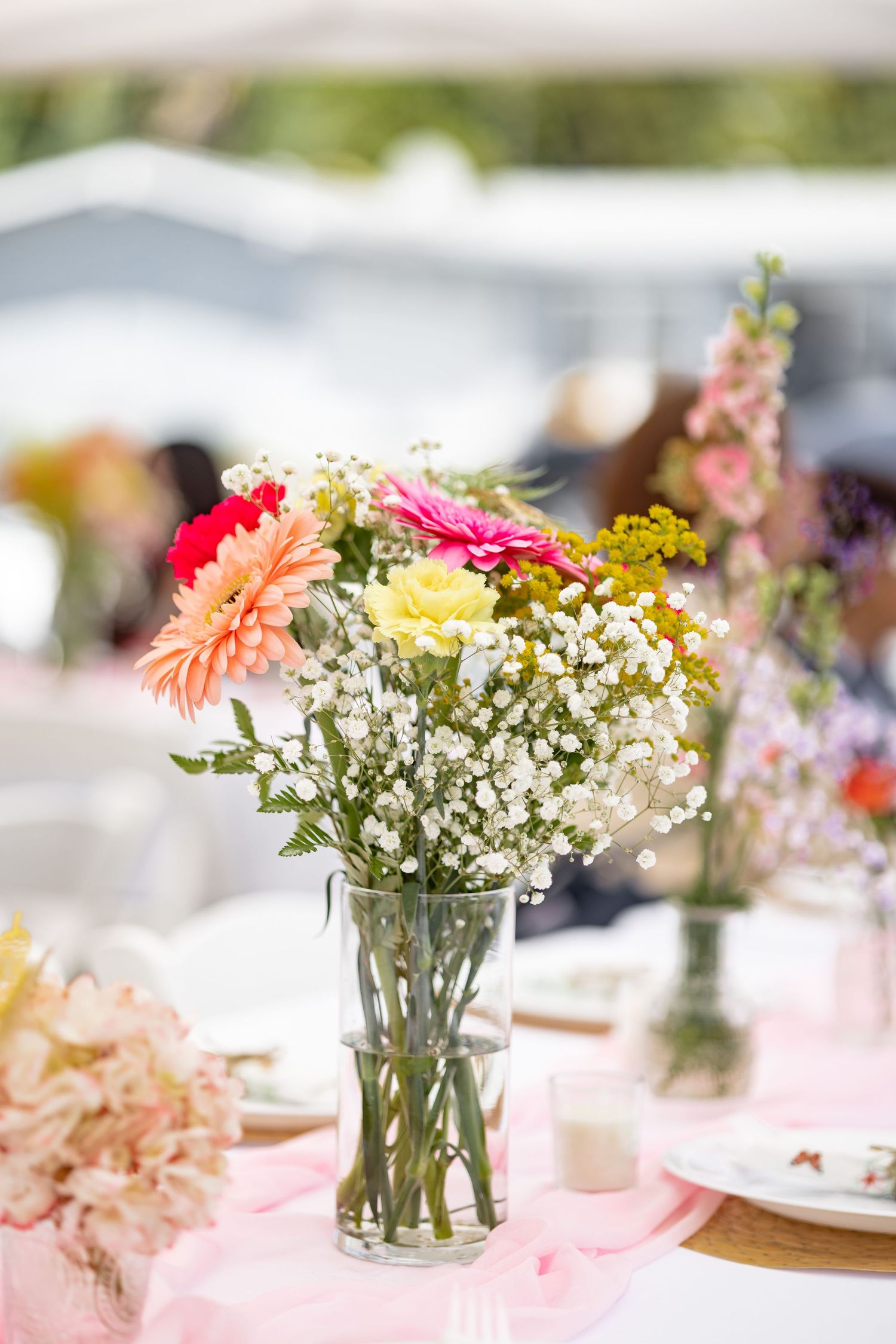 Colorful floral centerpiece with gerbera daisies and baby's breath in a clear glass vase on a pink tablecloth.