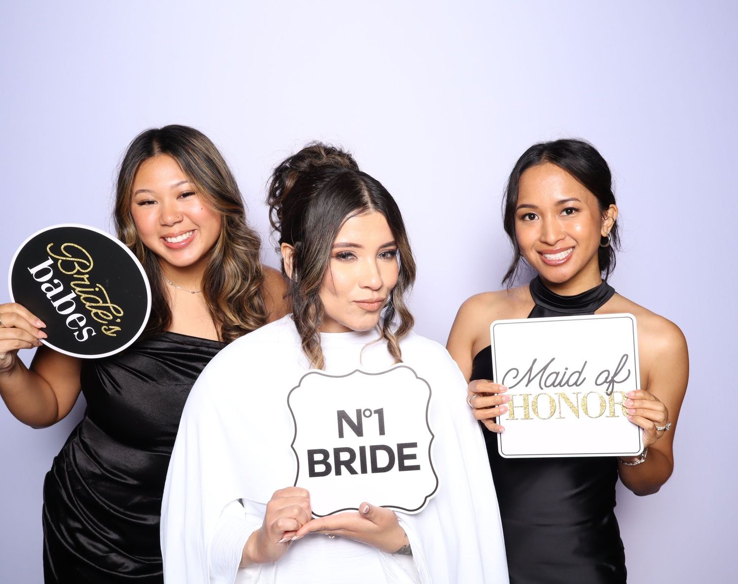 Three women pose together in a photo booth holding signs that say 'Bride' and 'Maid of Honor'.