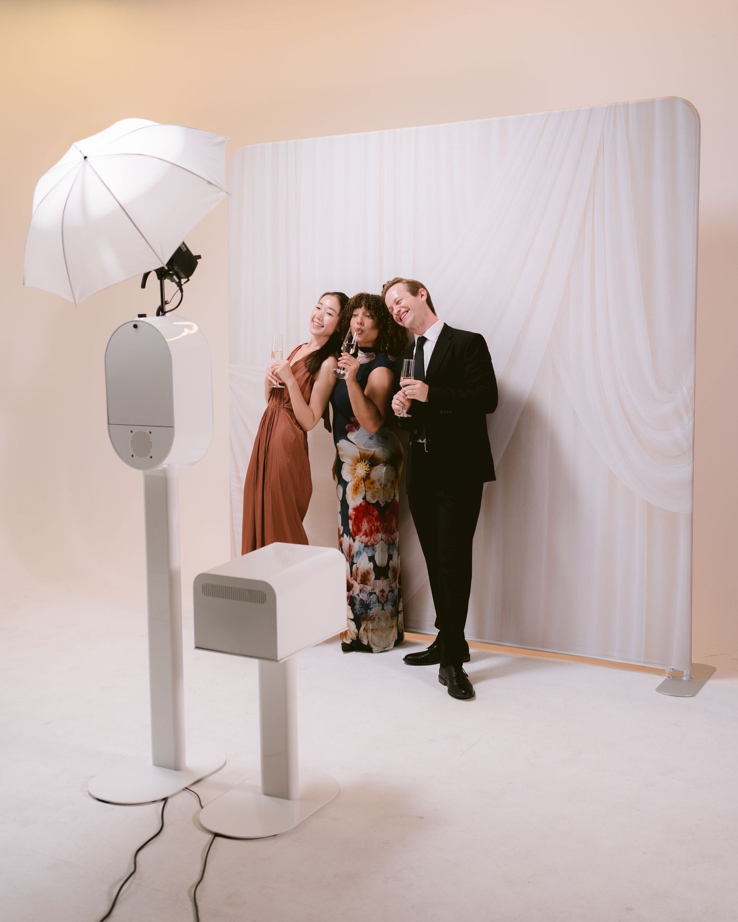 Three elegantly dressed guests pose and smile at a photo booth with white backdrop at a formal event.