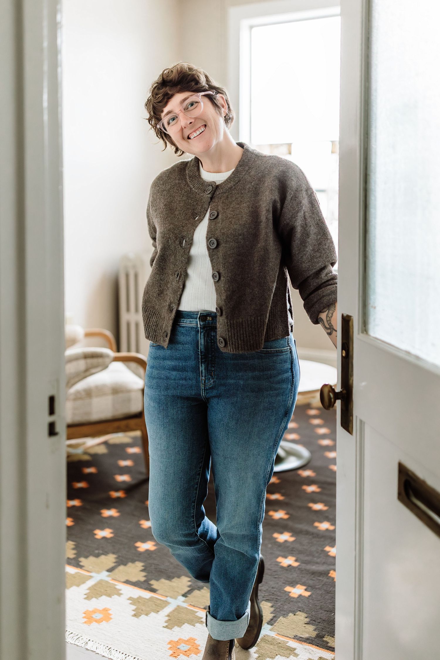 Cheerful woman in brown cardigan and jeans leaning against open white door in cozy home interior.