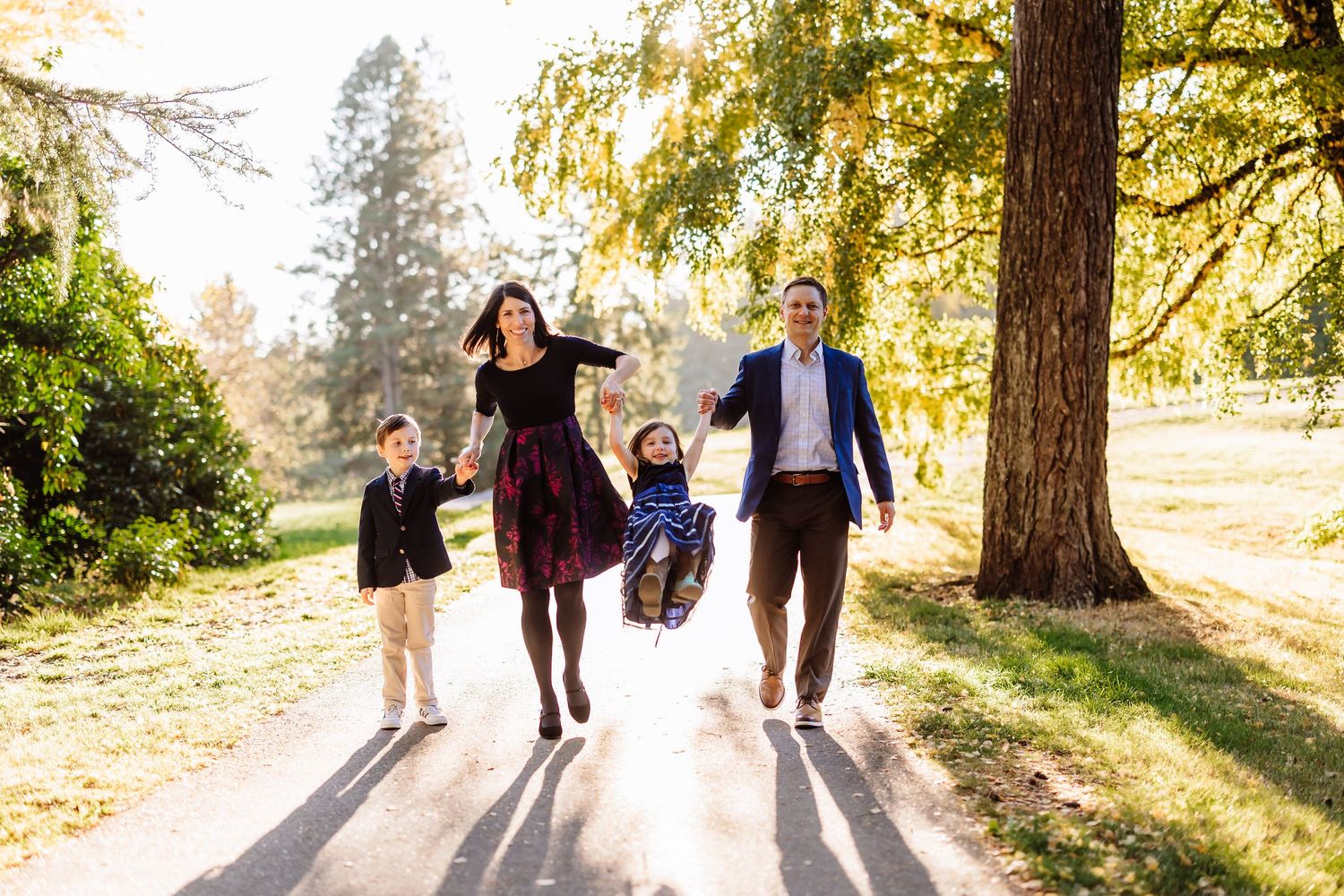 Family of four joyfully swinging child on sunny park path surrounded by golden autumn trees.