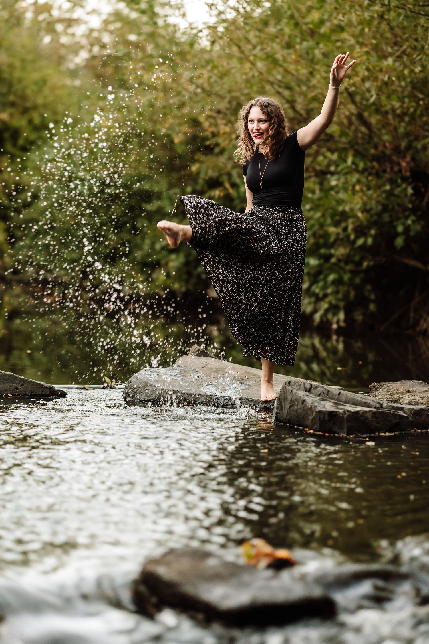 Woman in floral skirt joyfully splashing water on stepping stones in a lush green outdoor stream.