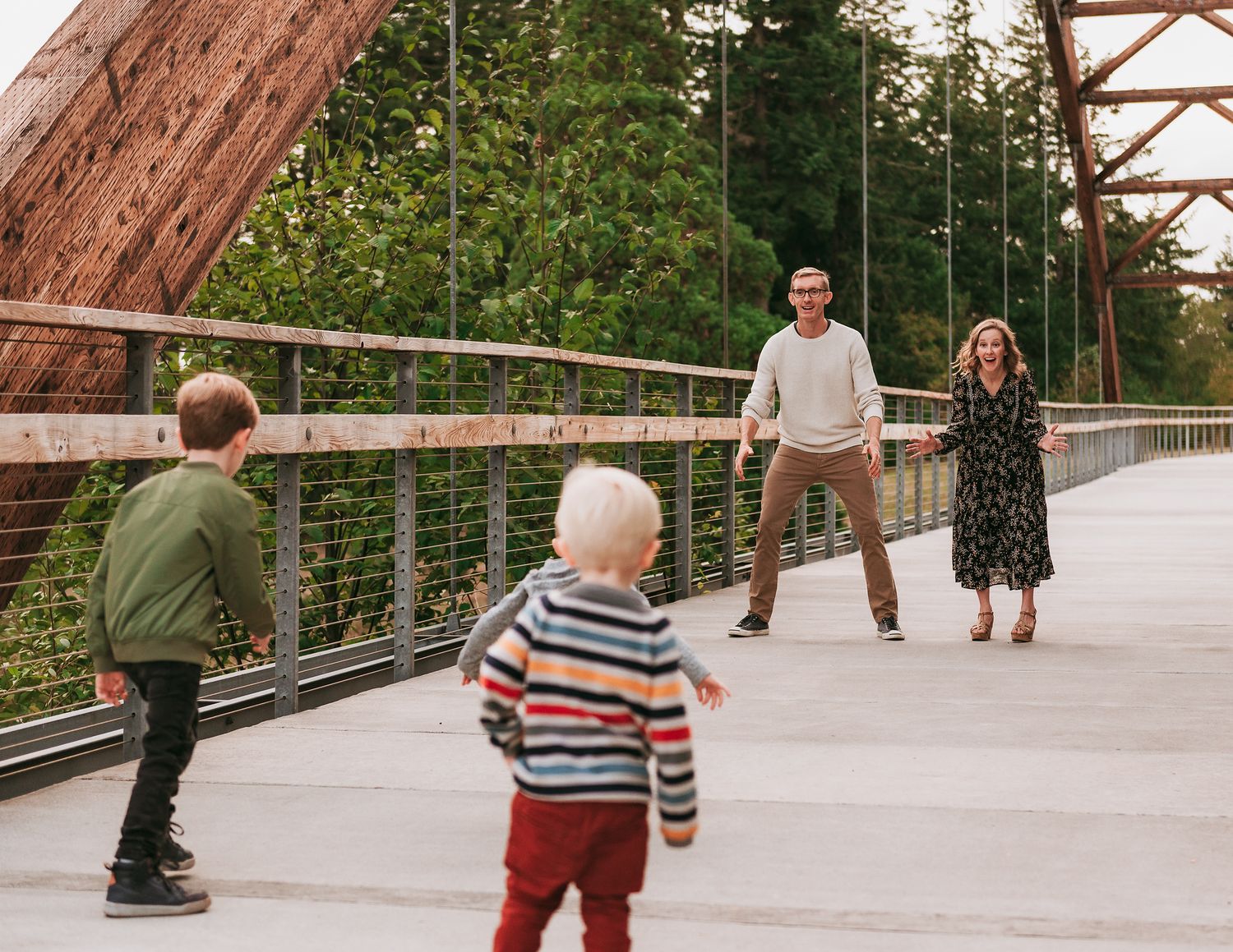 Family playing on a bridge with two young children, surrounded by lush green trees and modern railing.