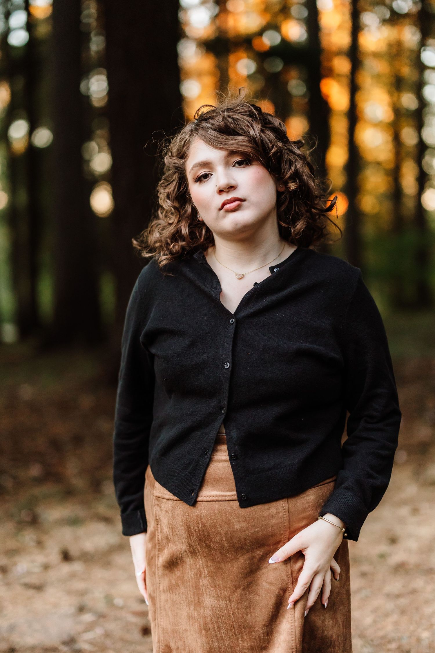 Young woman with curly hair in black cardigan and tan skirt posing in autumn forest with golden bokeh.