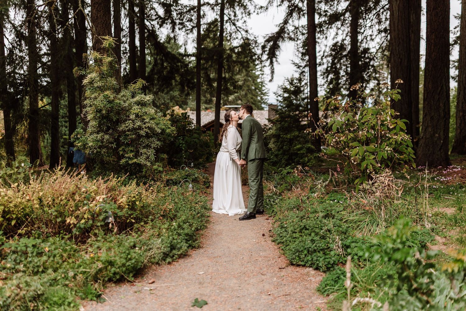 Couple embracing on a garden path surrounded by tall evergreen trees in a lush forest wedding setting.