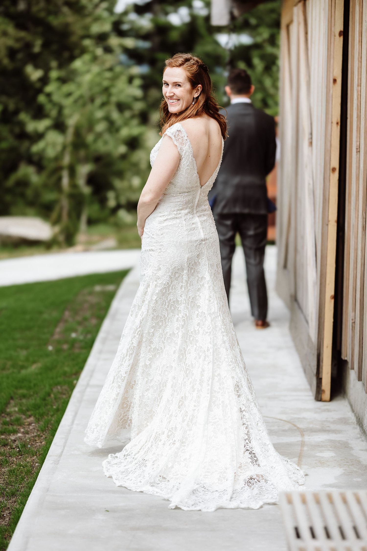 Bride in lace backless gown smiles over shoulder as groom walks ahead on pathway.