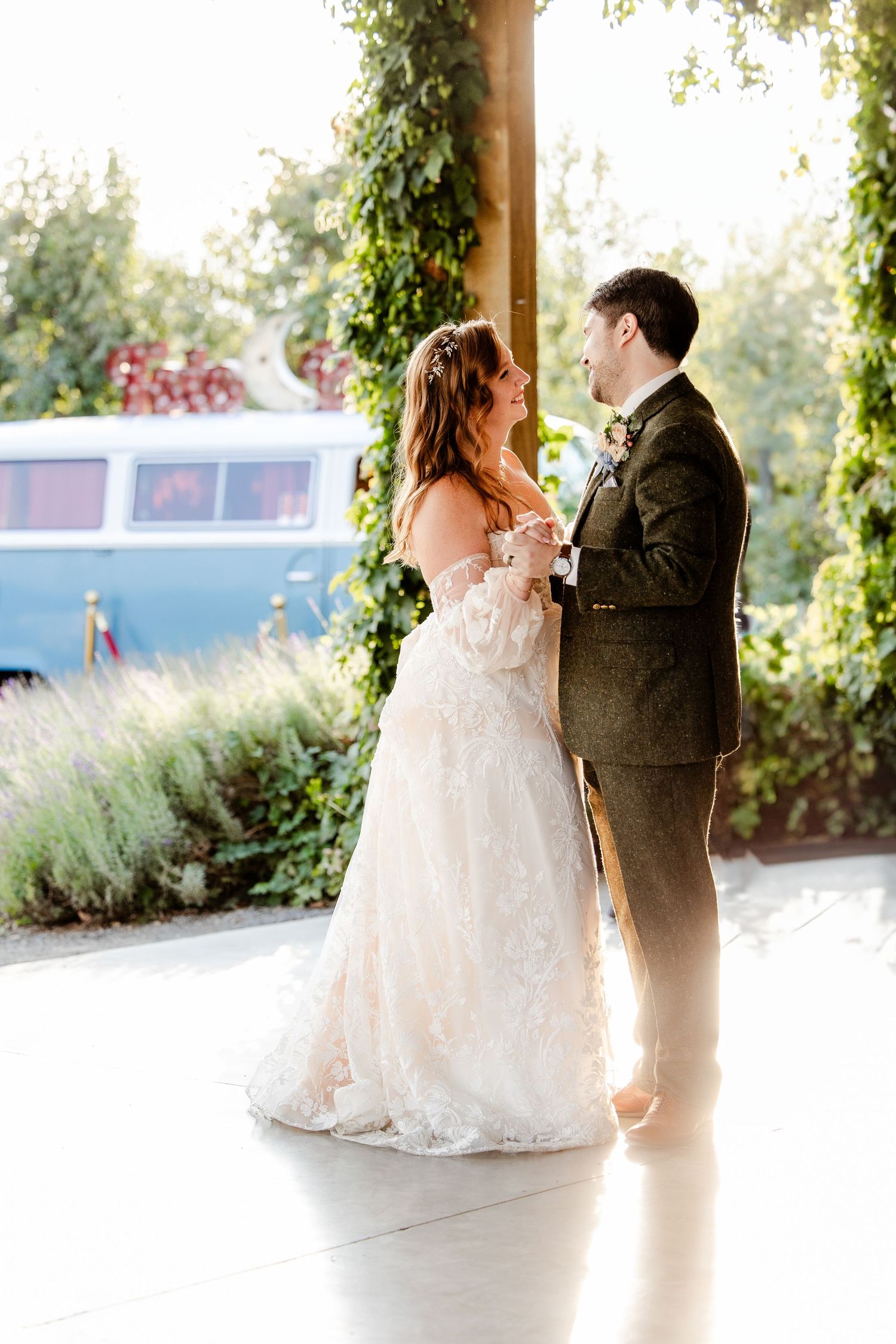 A bride and groom share their first dance on a sunlit dance floor with a vintage VW bus in the background.