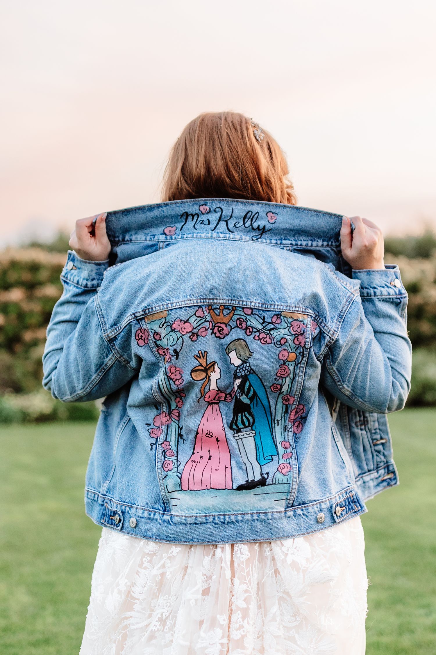 Bride displays custom painted denim jacket reading Mrs. Kelly with illustrated wedding couple design.