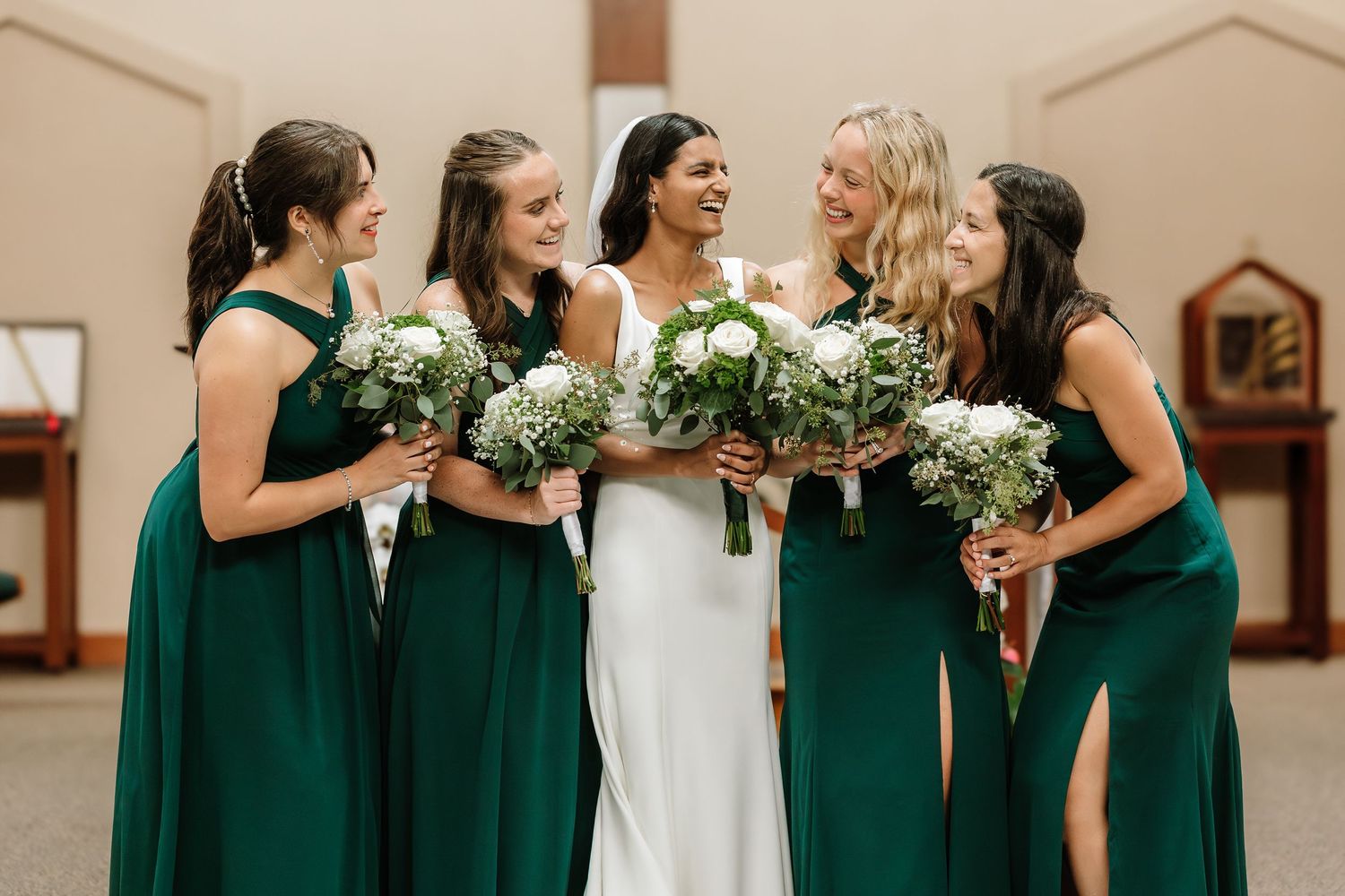 A bride in white stands with four bridesmaids in emerald green gowns holding white bouquets indoors.