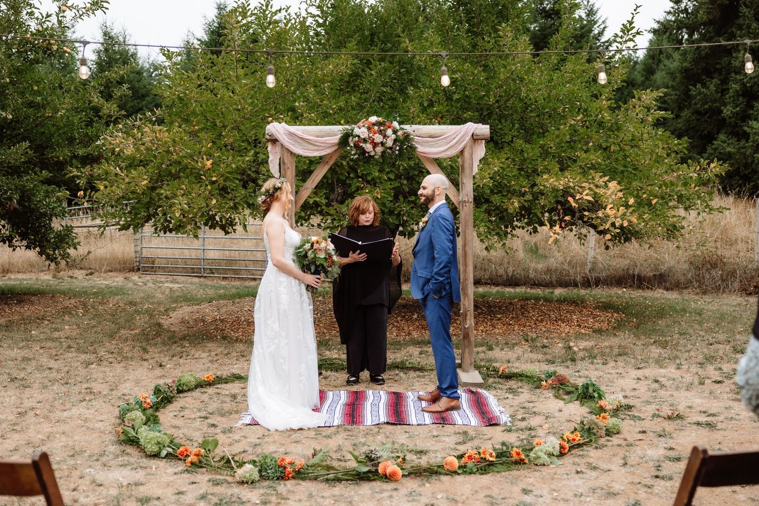 Outdoor boho wedding ceremony with couple at rustic arch, floral circle on ground, and officiant in natural setting.