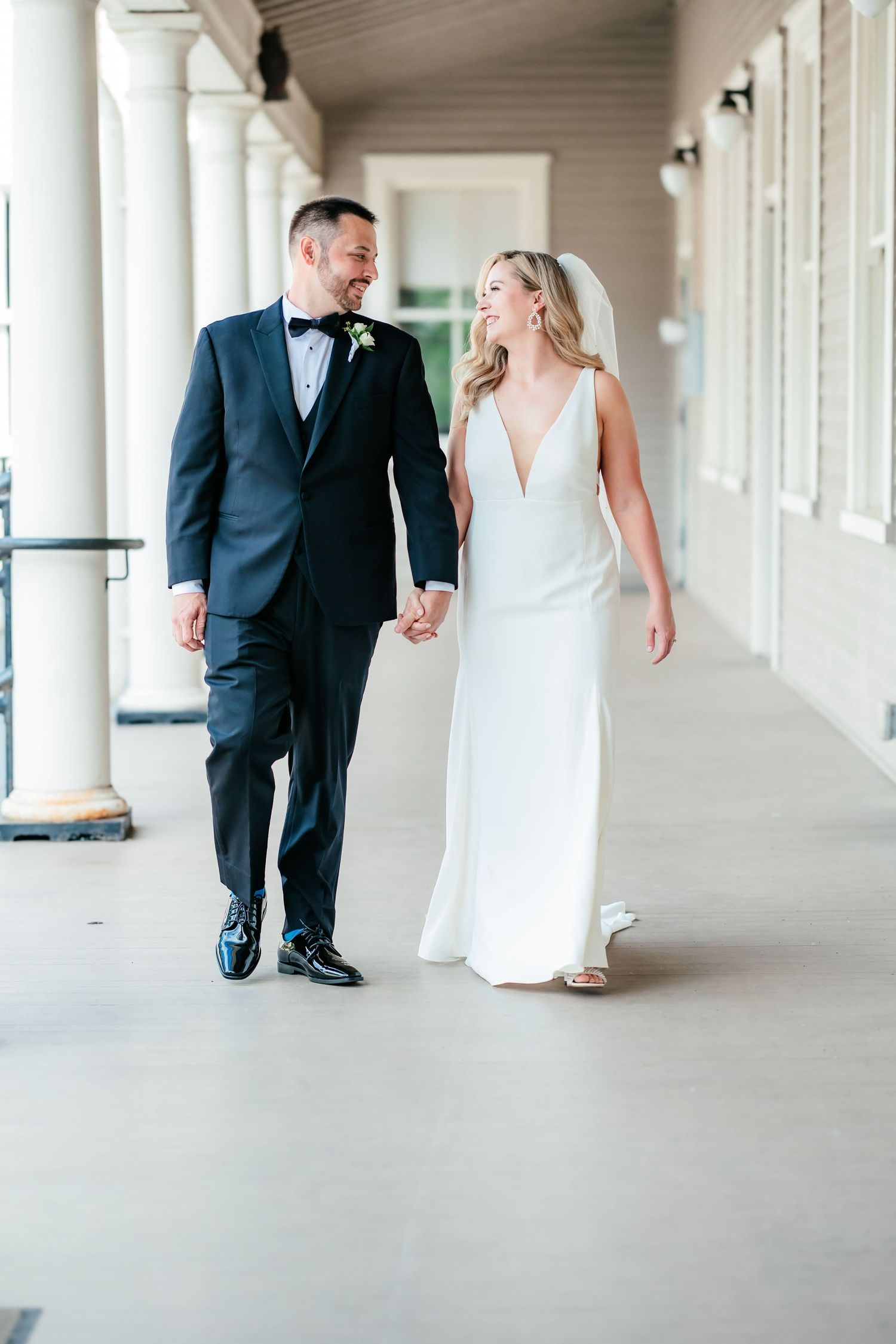 Bride and groom walking hand in hand along a grand columned outdoor corridor.