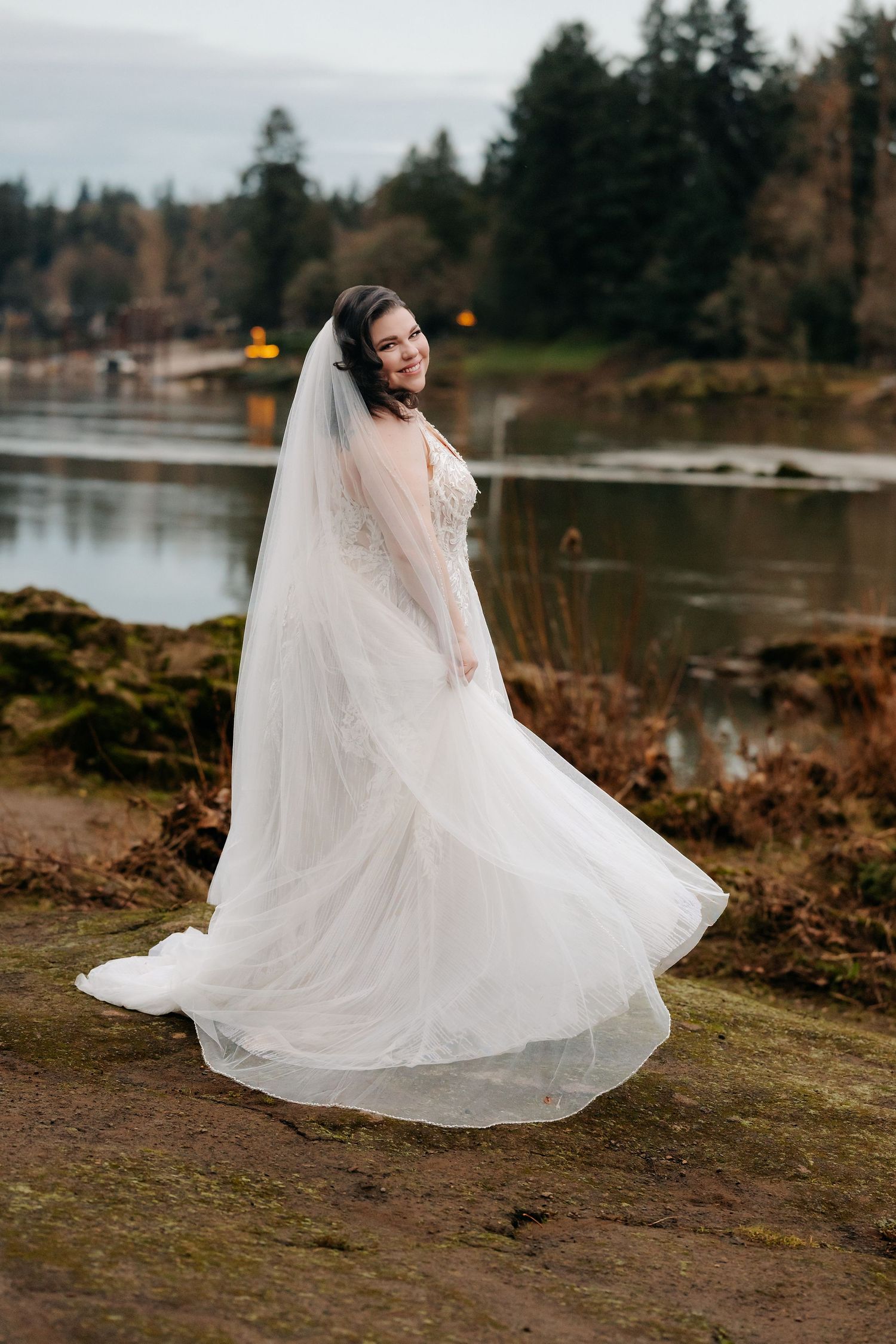 Bride twirling in a flowing white gown and veil beside a serene lake at dusk.