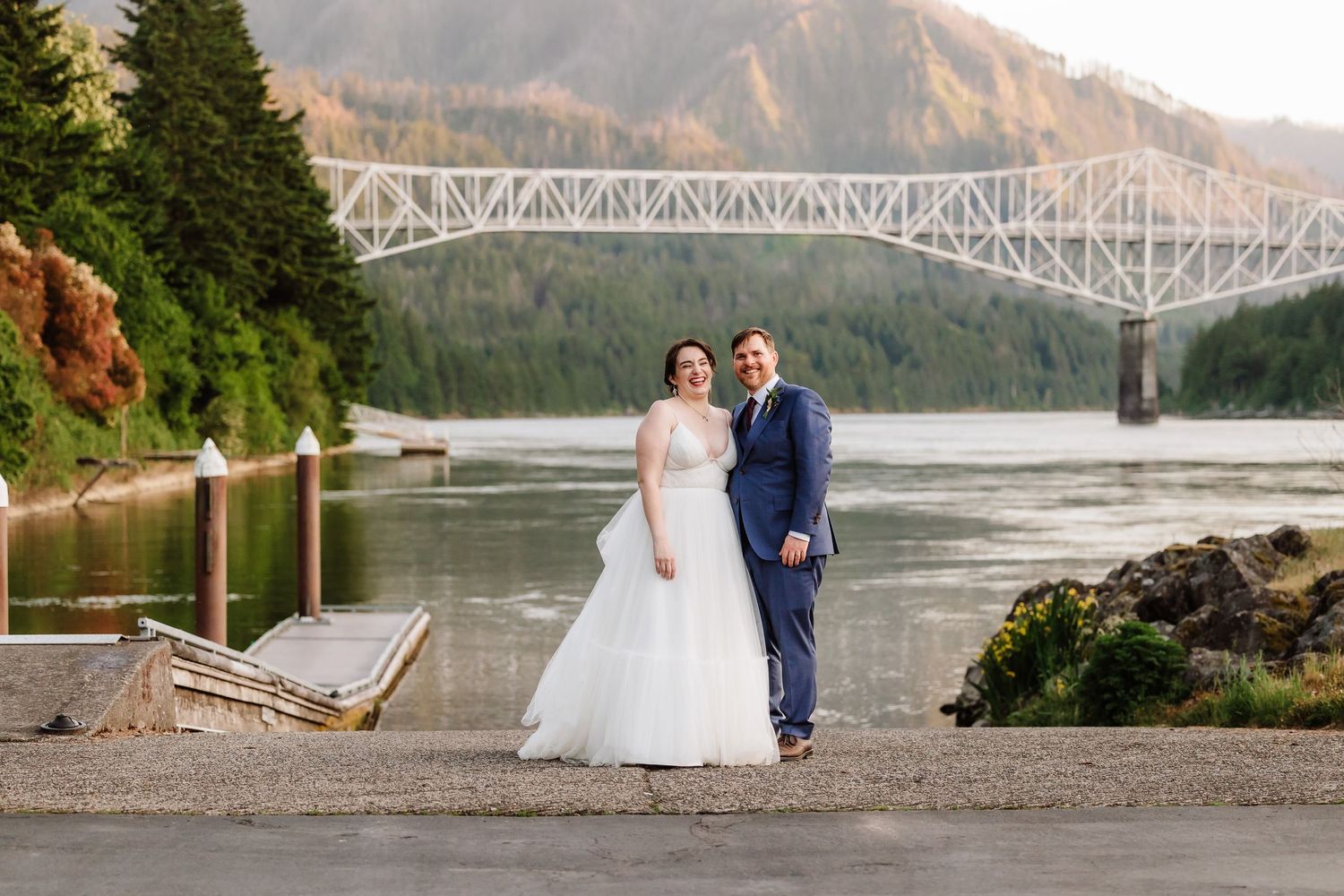 Smiling bride and groom pose by a river with a large bridge and autumn mountains behind them.