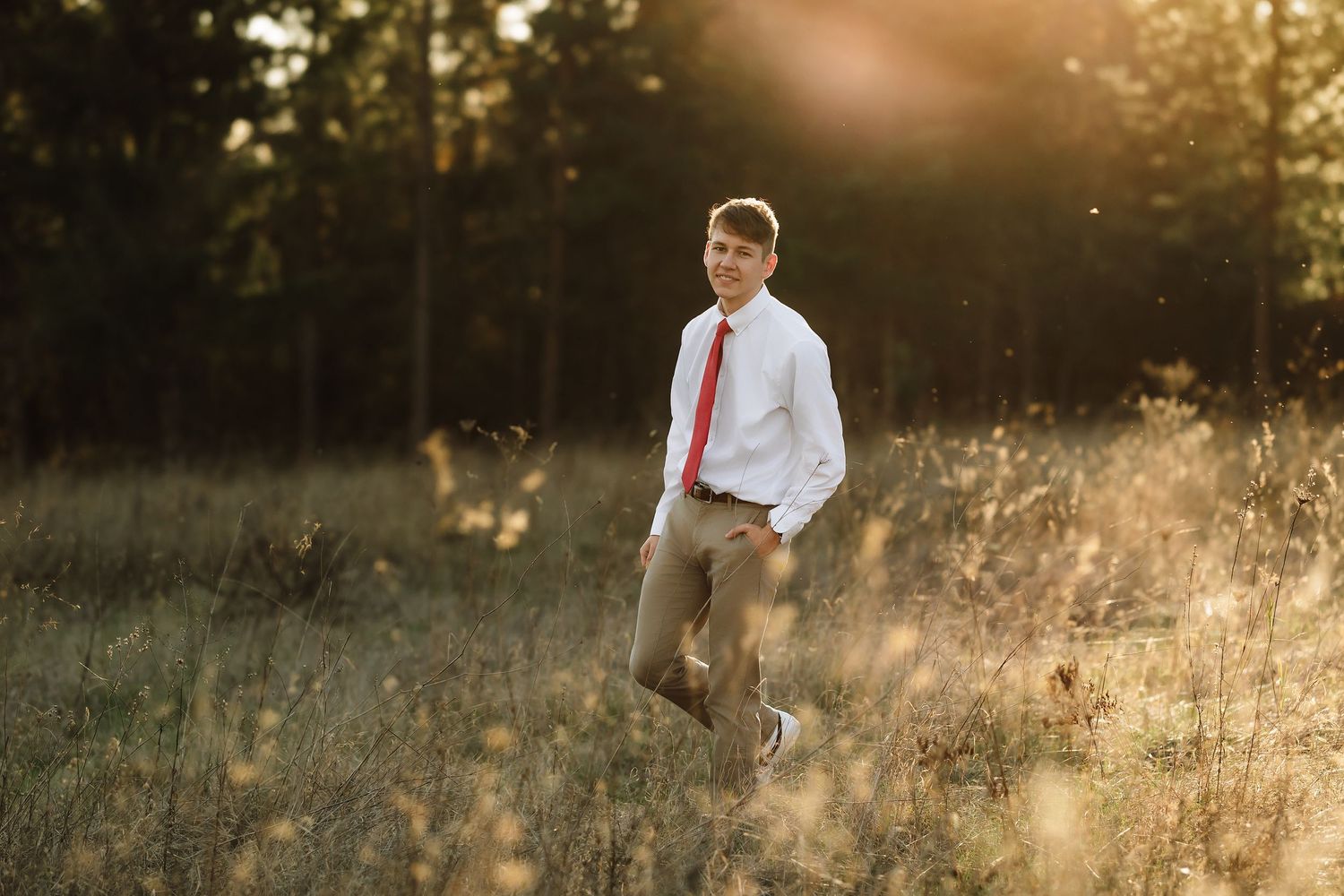 A young boy in a white shirt and red tie poses in a sunlit field with golden hour light and trees in the background.