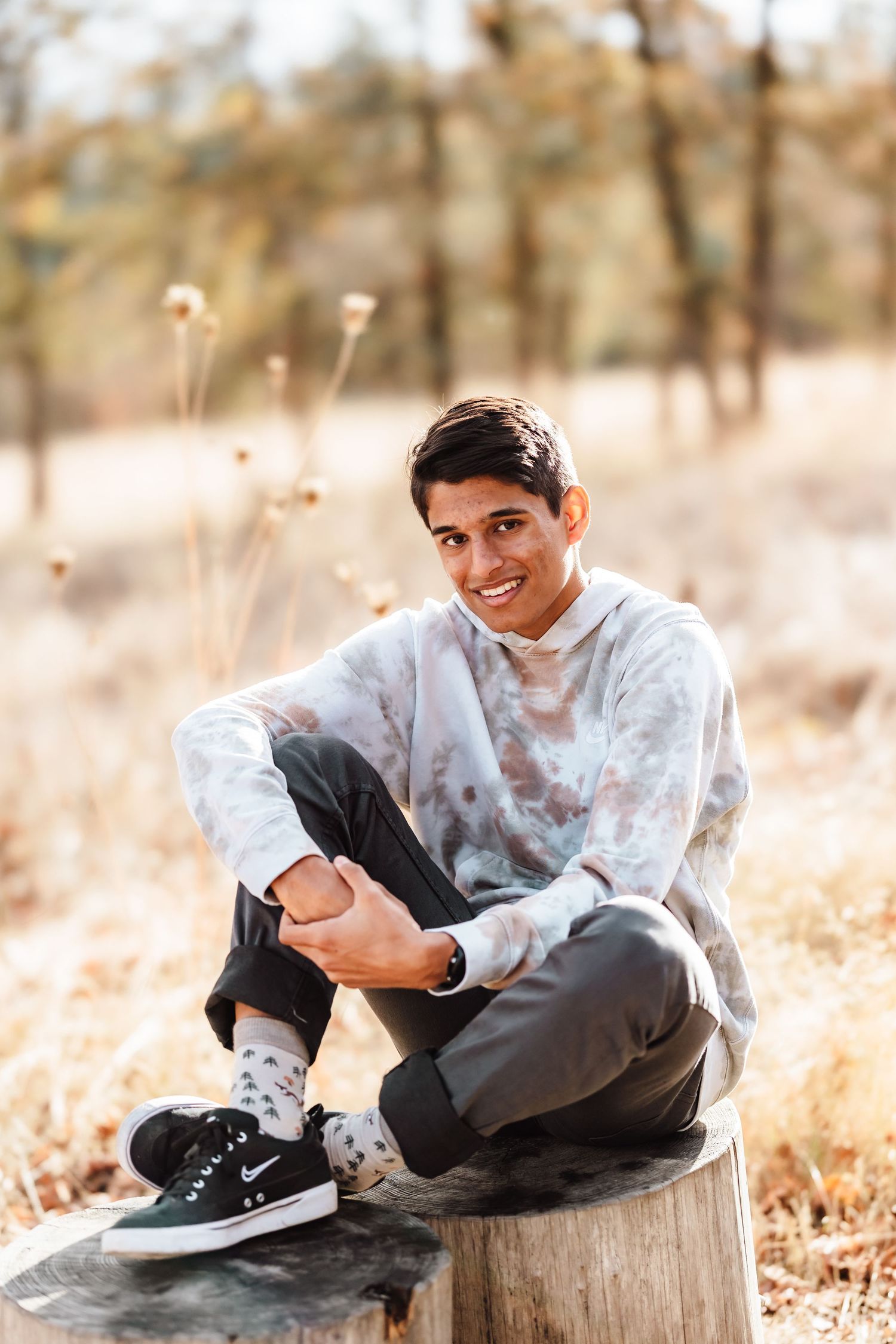 Young man smiling, sitting on a tree stump outdoors in fall, wearing tie-dye sweatshirt, gray pants, and Nike sneakers.