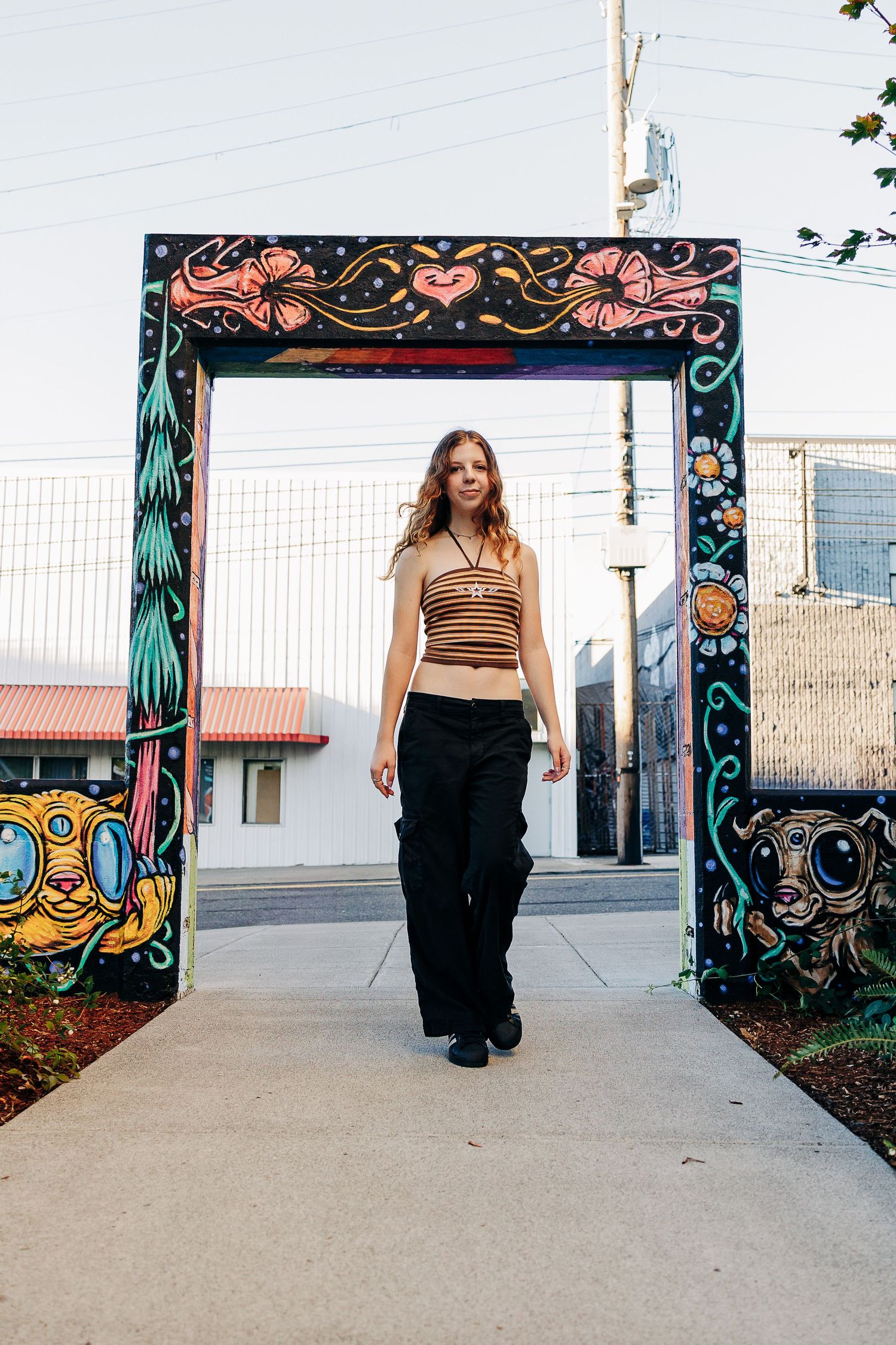Woman in striped crop top and black wide-leg pants walking through colorful graffiti art arch outdoors.