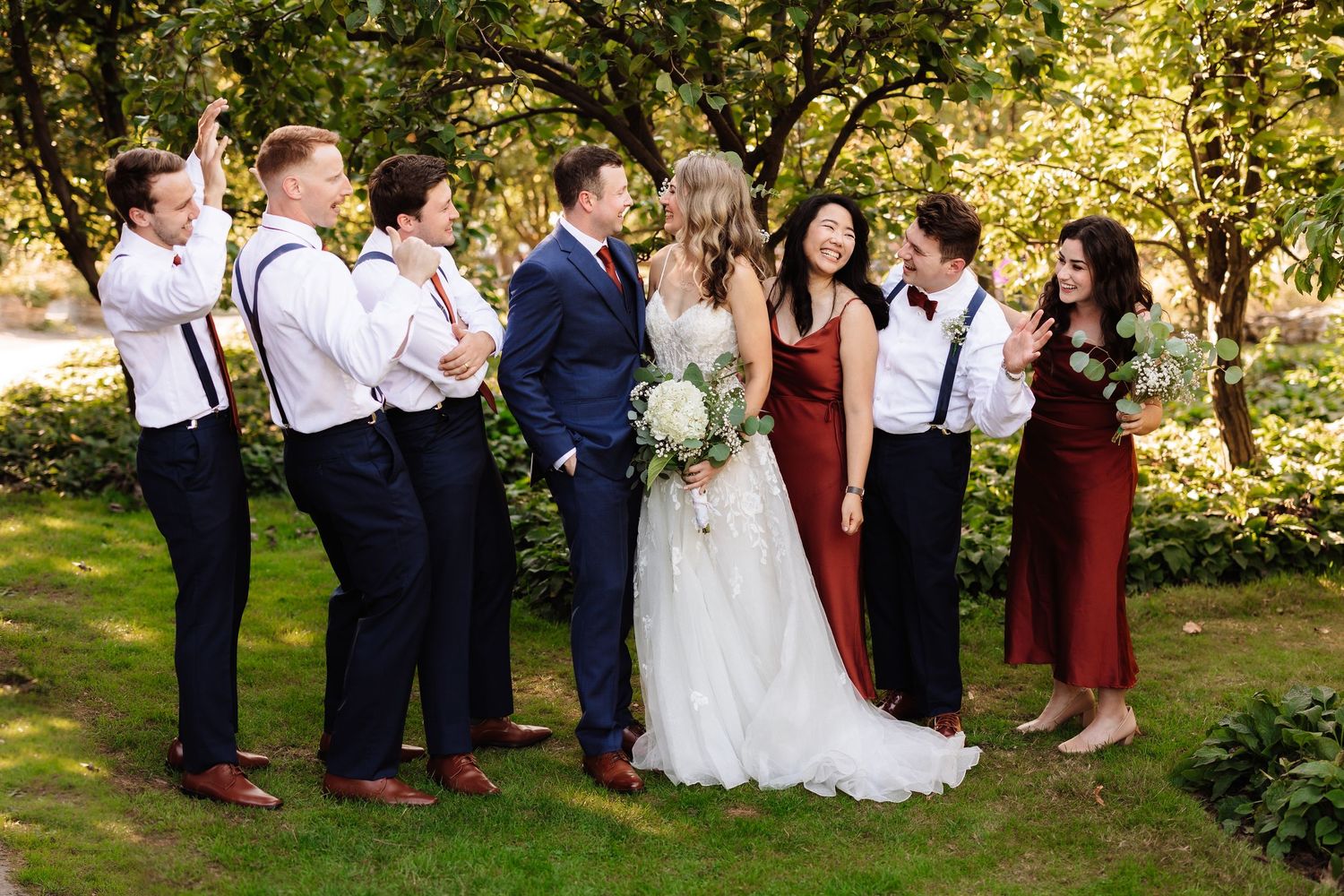 Wedding party laughing together outdoors with bride and groom kissing in the center.