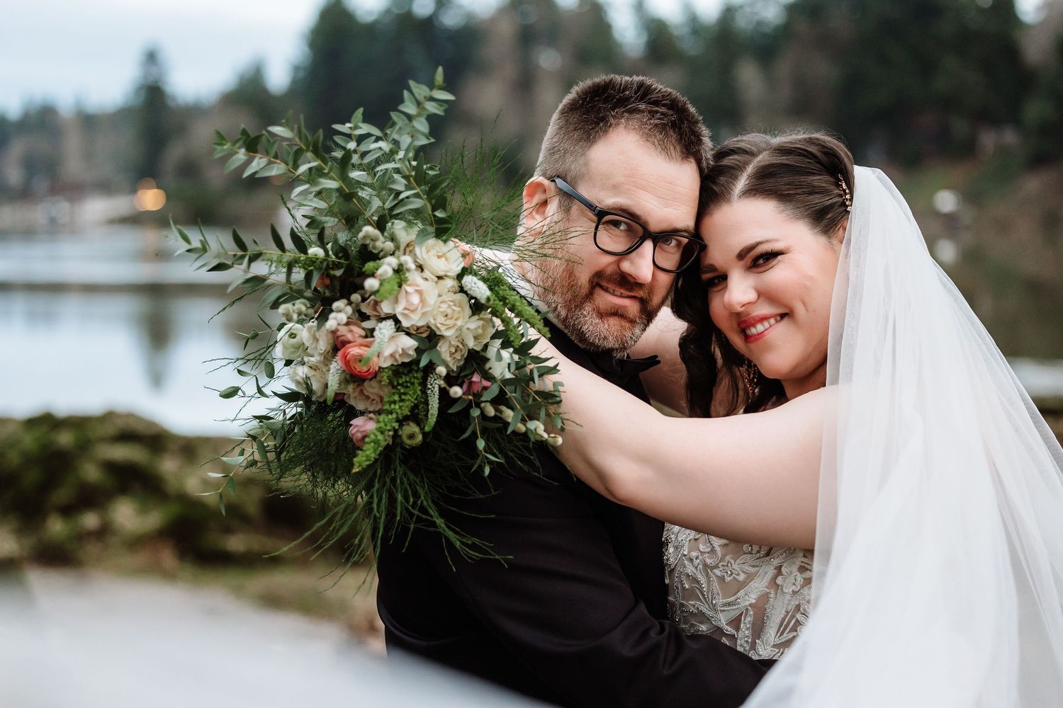 Bride and groom embracing with floral bouquet near a lakeside venue on wedding day.