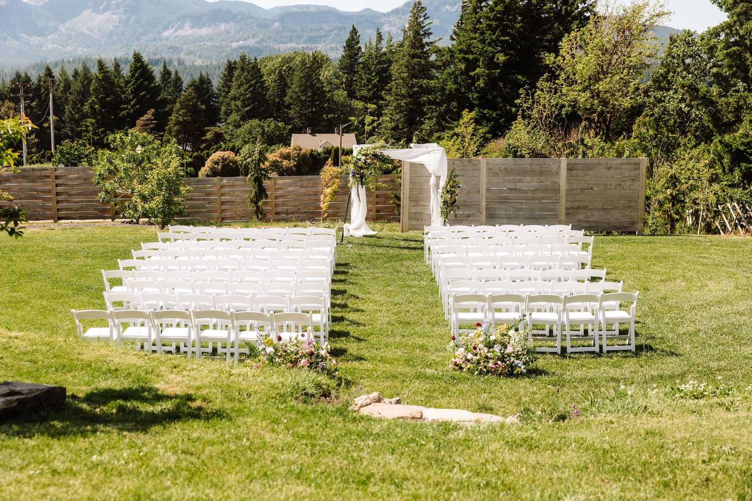 Outdoor wedding ceremony setup with white folding chairs on green lawn with floral arch and mountain views.