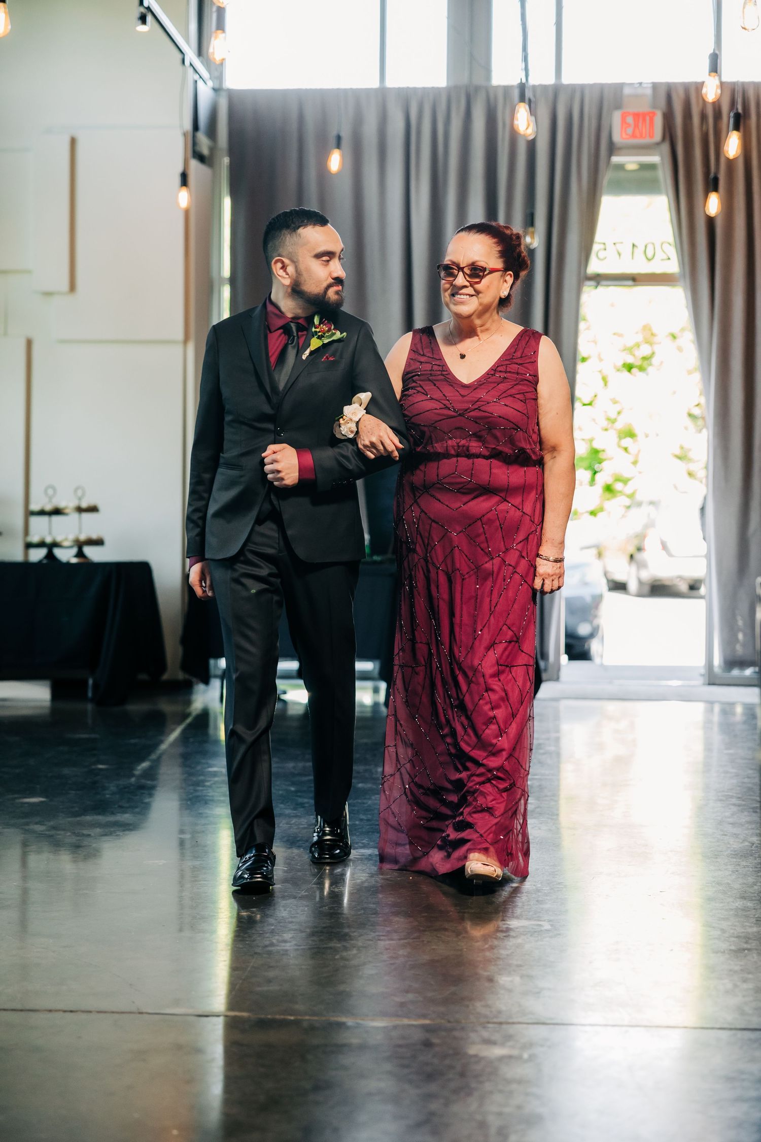 Groom in black suit escorts mother in burgundy gown down wedding ceremony aisle.