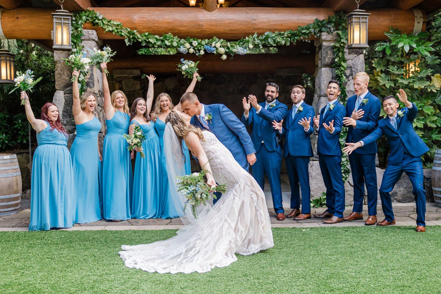 Wedding party celebrating as bride and groom kiss, bridesmaids in blue and groomsmen in navy suits.