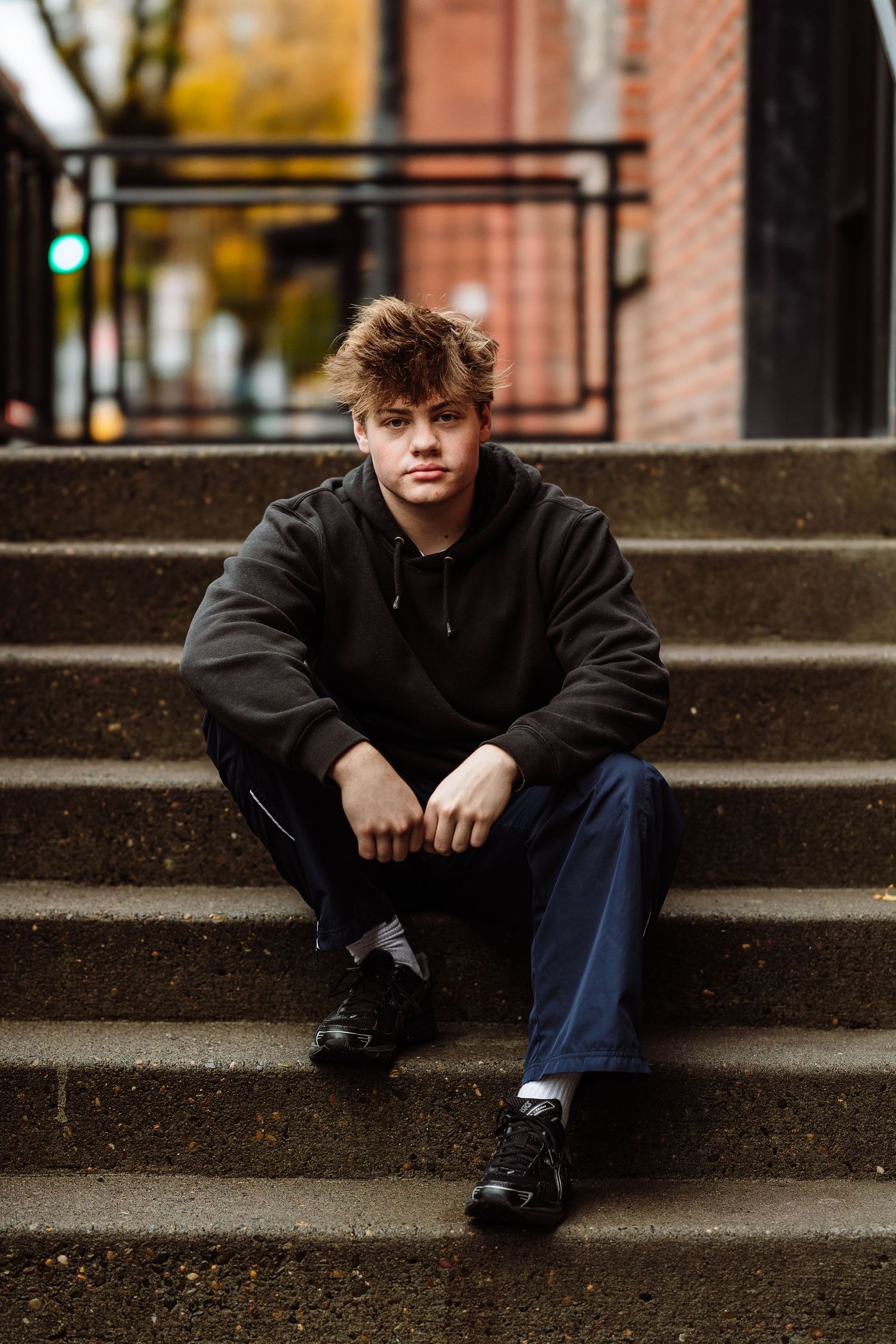 Young man in black hoodie and jeans sitting on concrete steps outdoors in autumn.