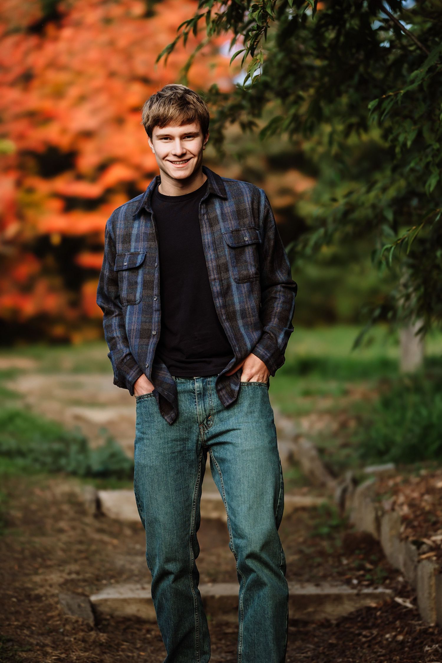 Young man in plaid shirt and jeans smiling outdoors with vibrant autumn foliage in background.