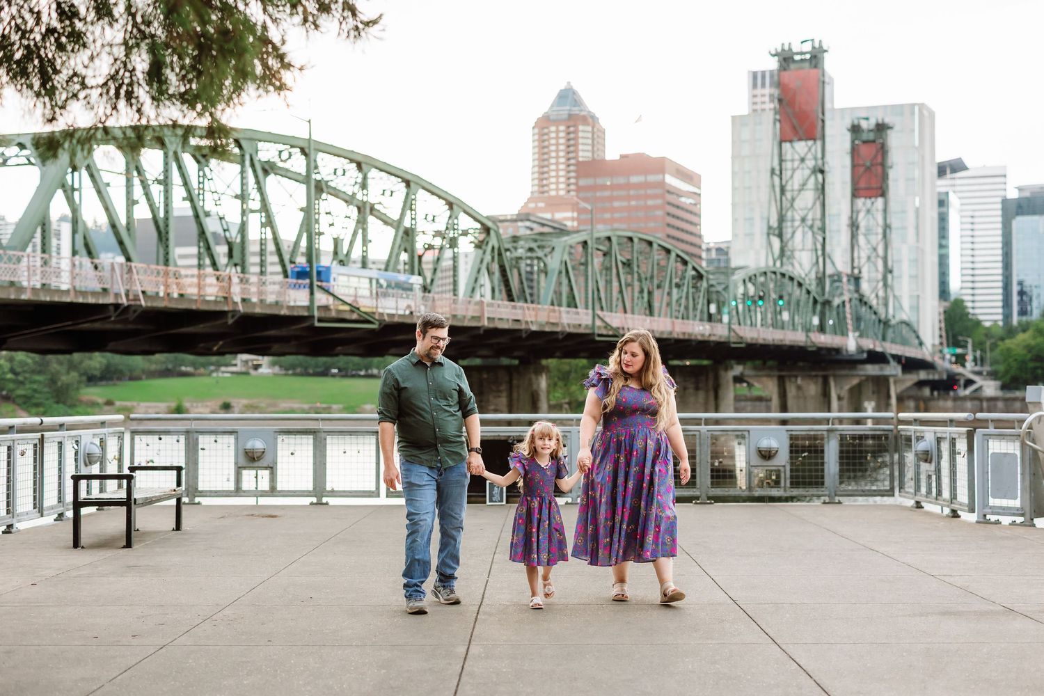 Family of three walking hand in hand on waterfront promenade with Portland city bridge and skyline in background.