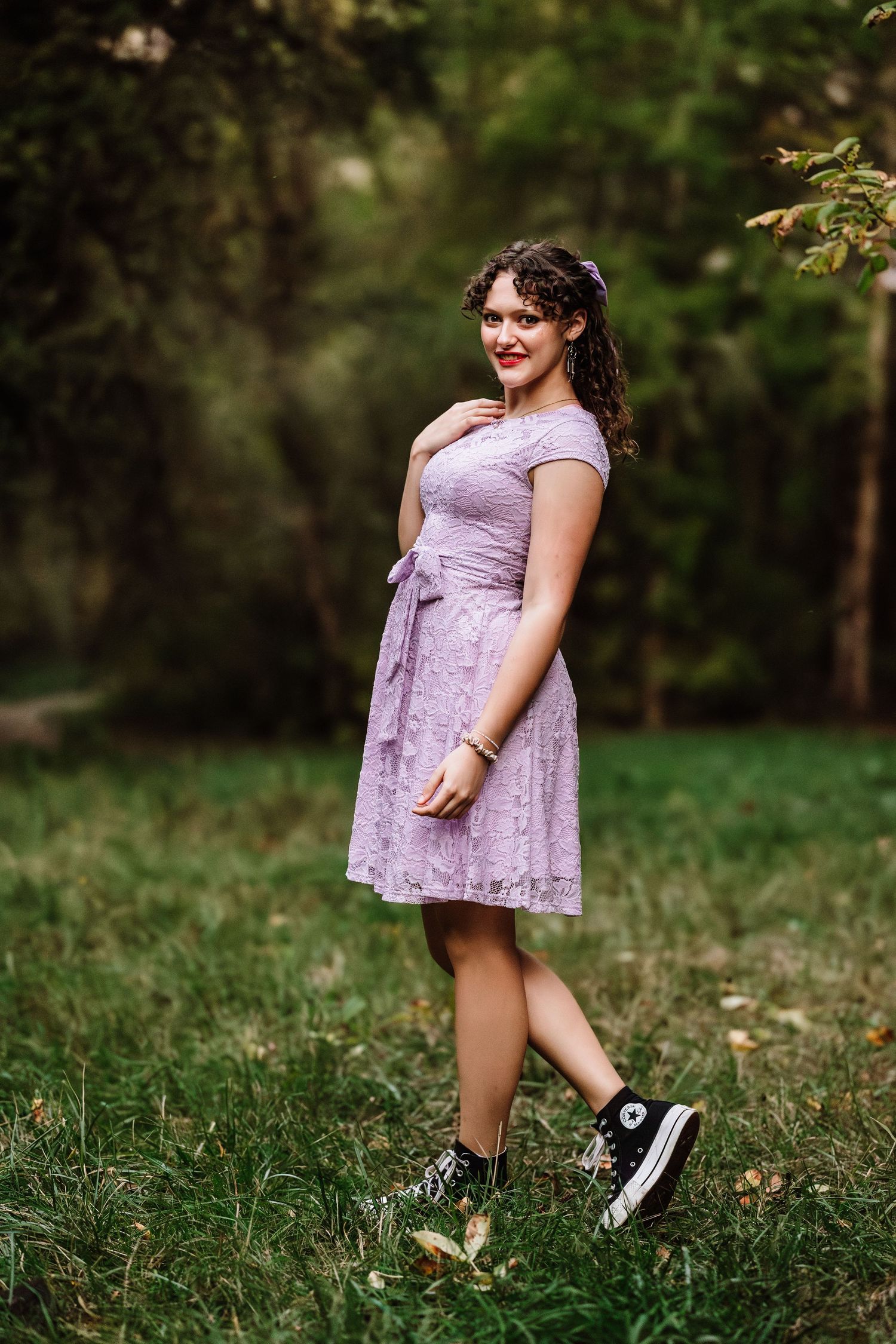 Young woman in lavender lace dress and black Converse sneakers posing outdoors in a green park setting.