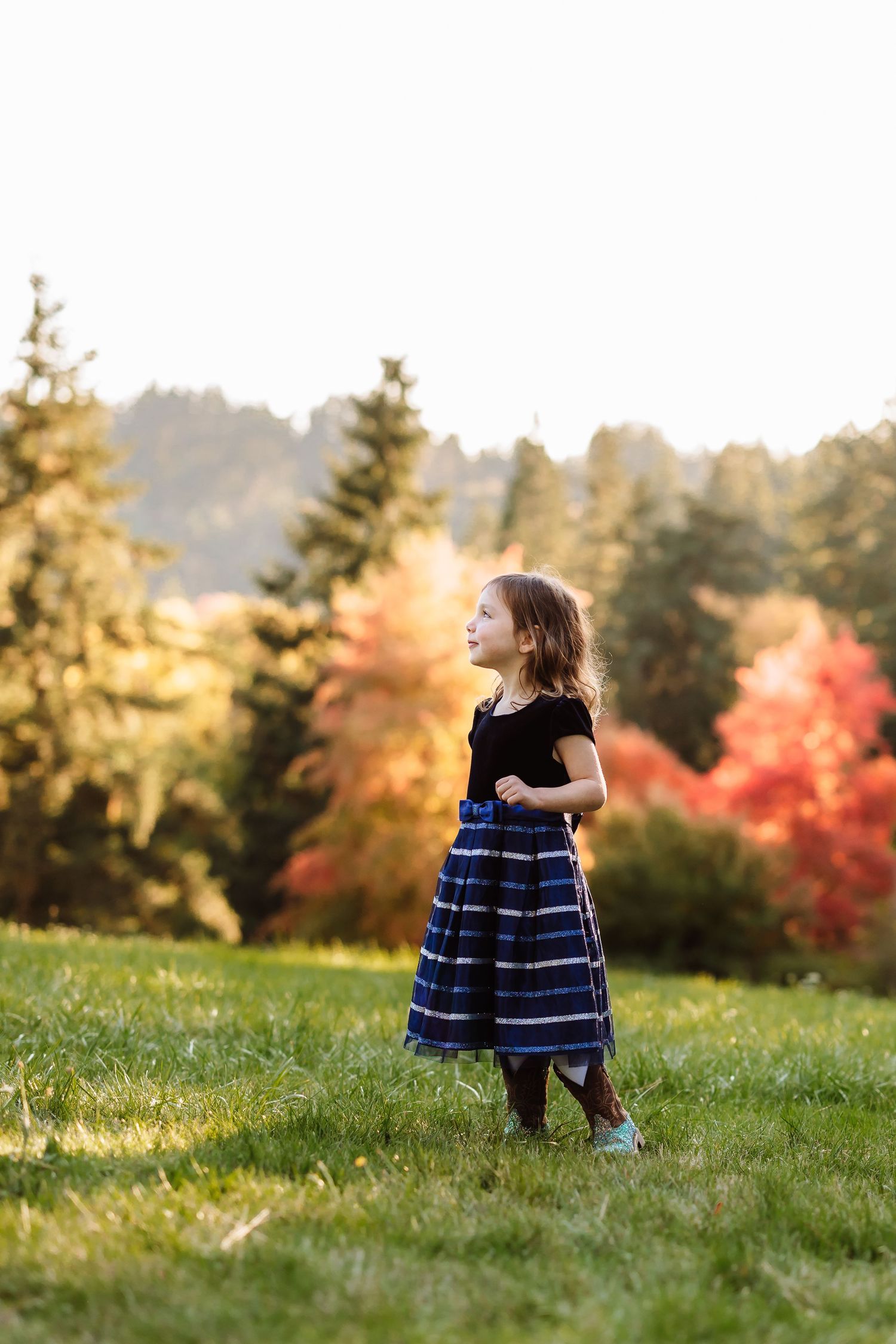 Young girl in black top and blue plaid skirt standing on green grass with colorful autumn trees in background.