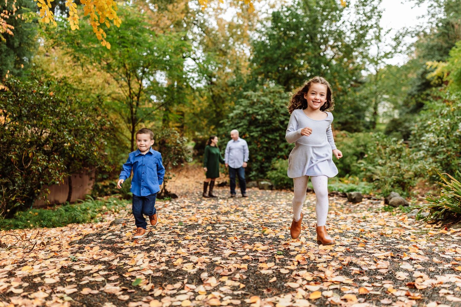Two children run joyfully on a leaf-covered path in an autumn park with family in background.