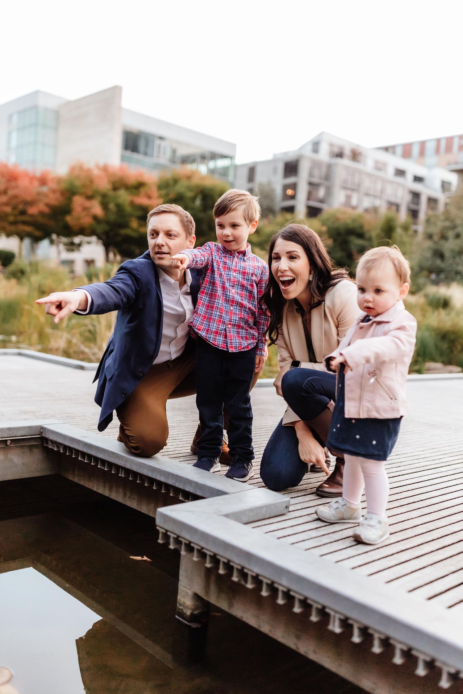 Family of four posing on a wooden dock, pointing excitedly, with urban buildings and autumn trees in the background.