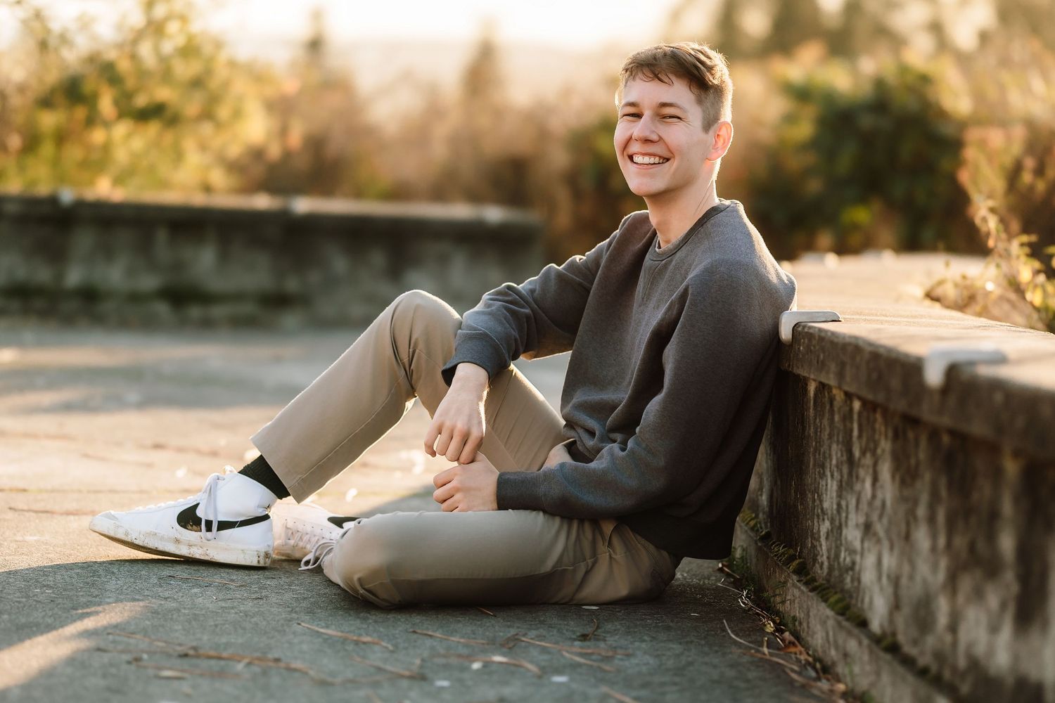 Young man smiling, sitting against a stone wall outdoors in casual khaki pants and gray sweater with white sneakers.