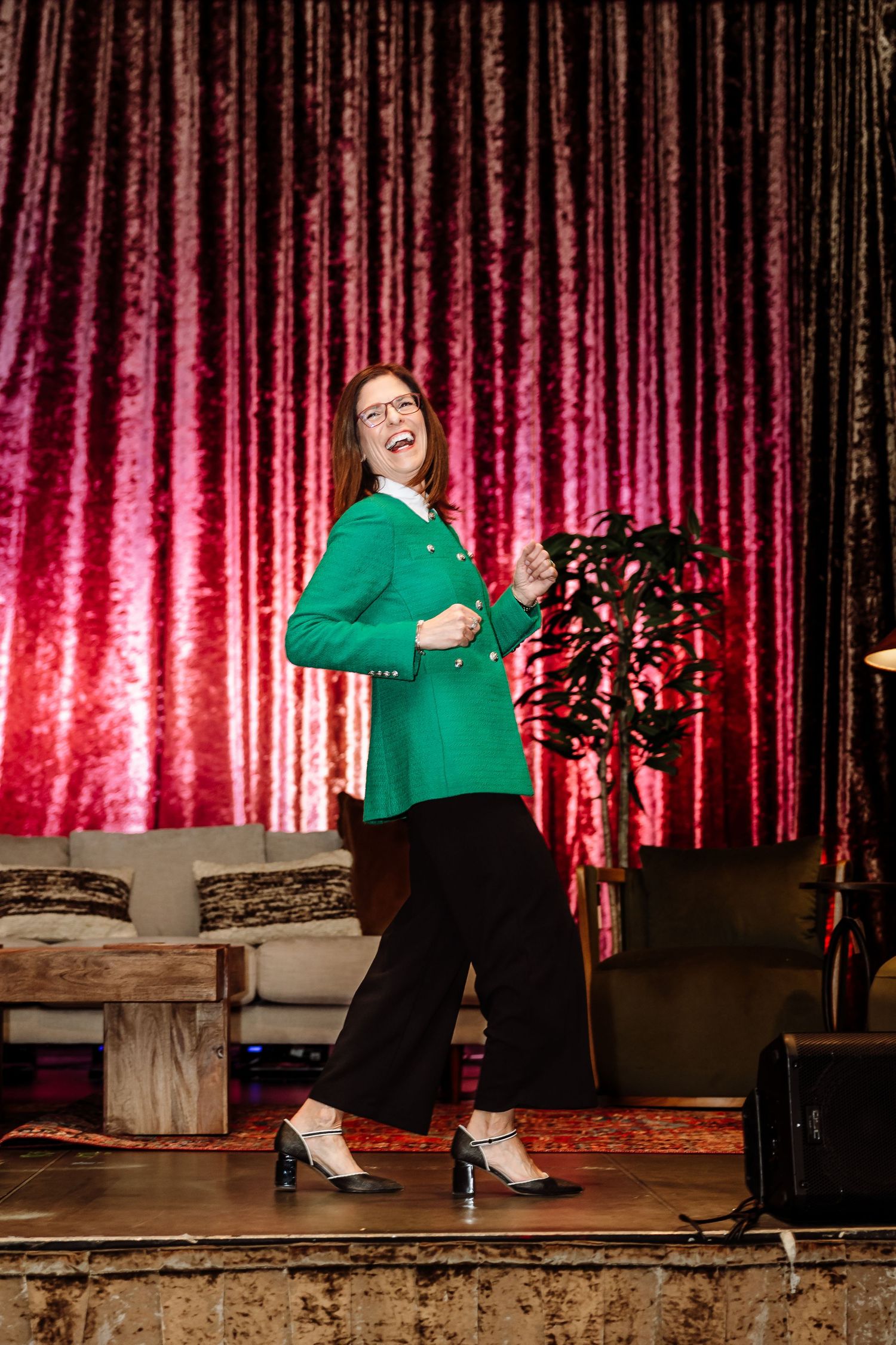 Woman in green blazer laughing on stage in front of a red velvet curtain backdrop.