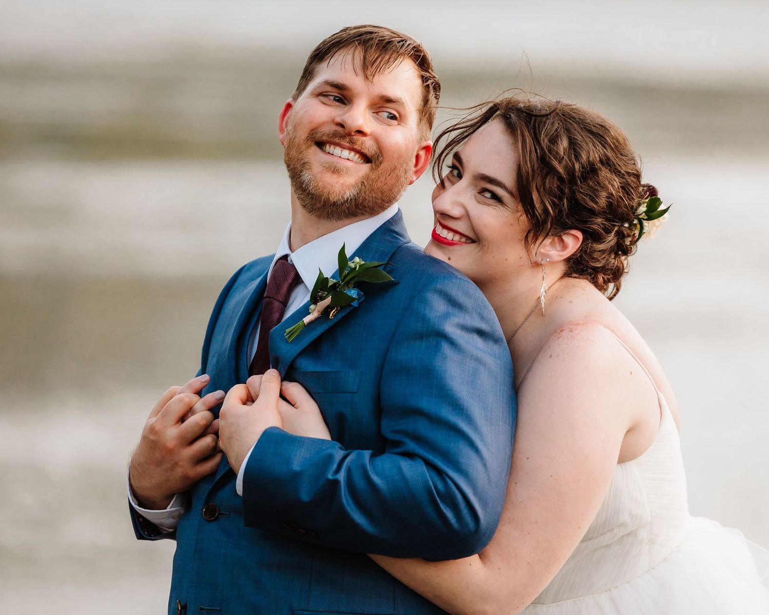 Joyful bride hugging groom from behind, both smiling, groom in blue suit with floral boutonniere.