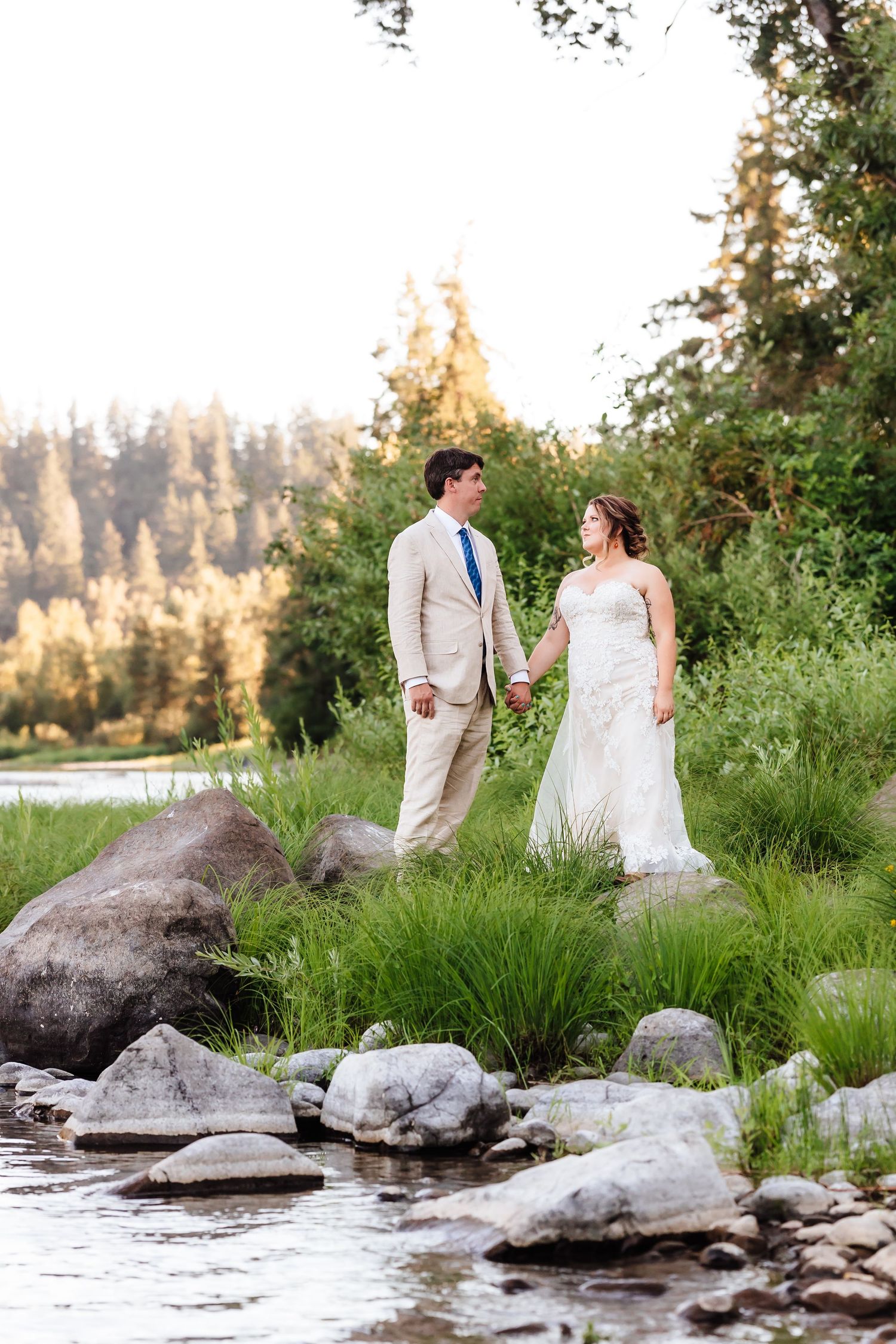 Bride and groom holding hands on a rocky riverbank with tall green grass and a sunlit forest in the background.