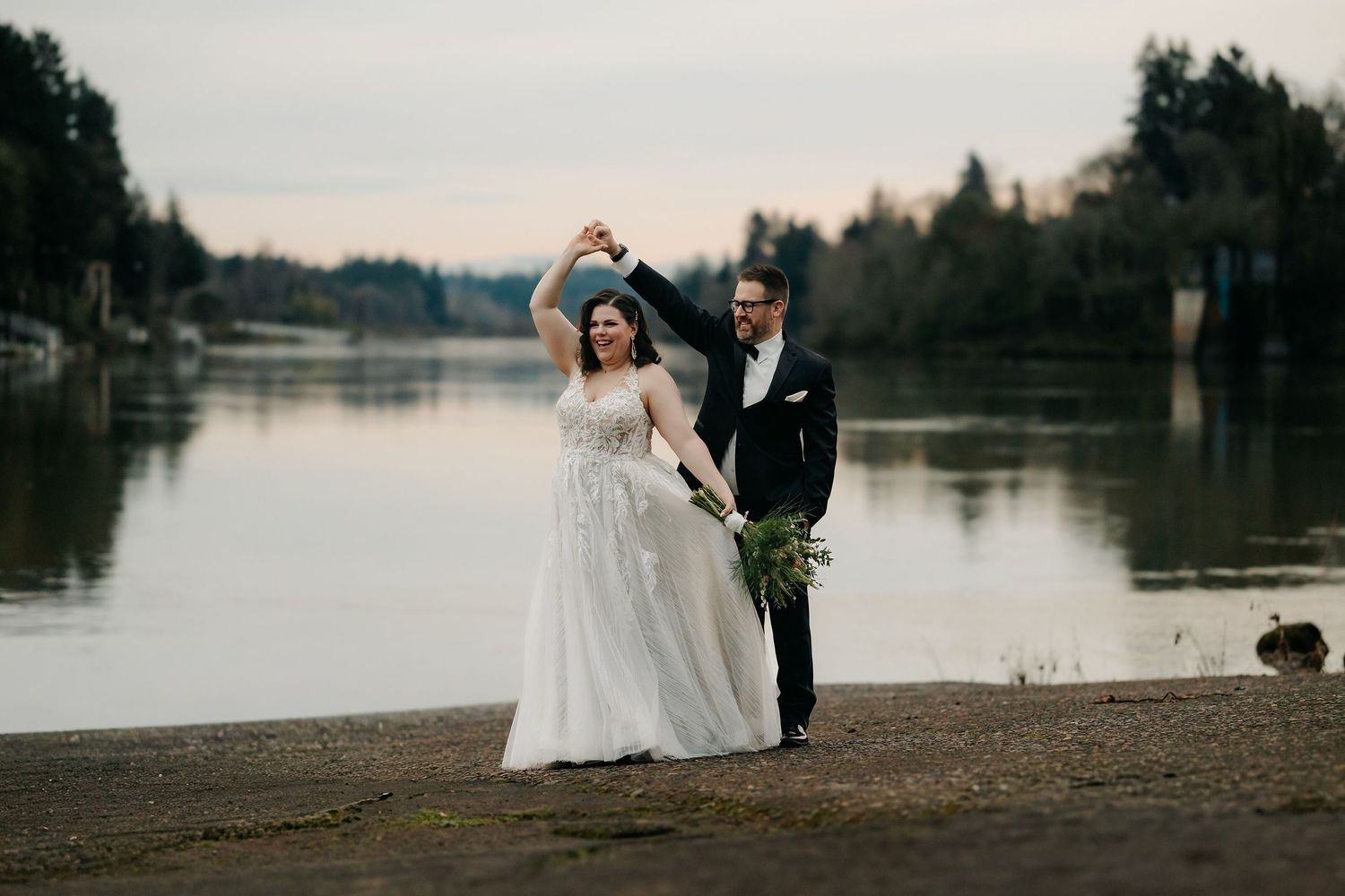Bride and groom dancing by a serene lake at dusk, bride in flowing white gown holding bouquet.