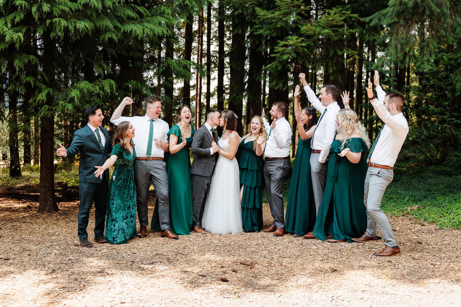 Bridal party celebrates in forest, bridesmaids in emerald green dresses, groomsmen in white shirts and gray pants.