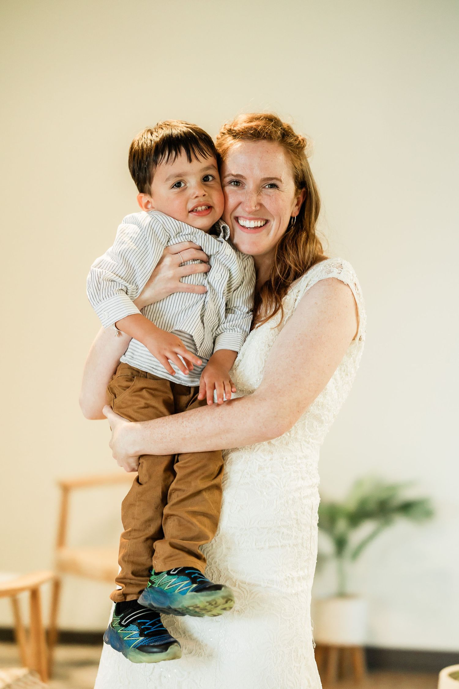 Smiling bride in white dress holds young toddler boy at bright and airy wedding reception.