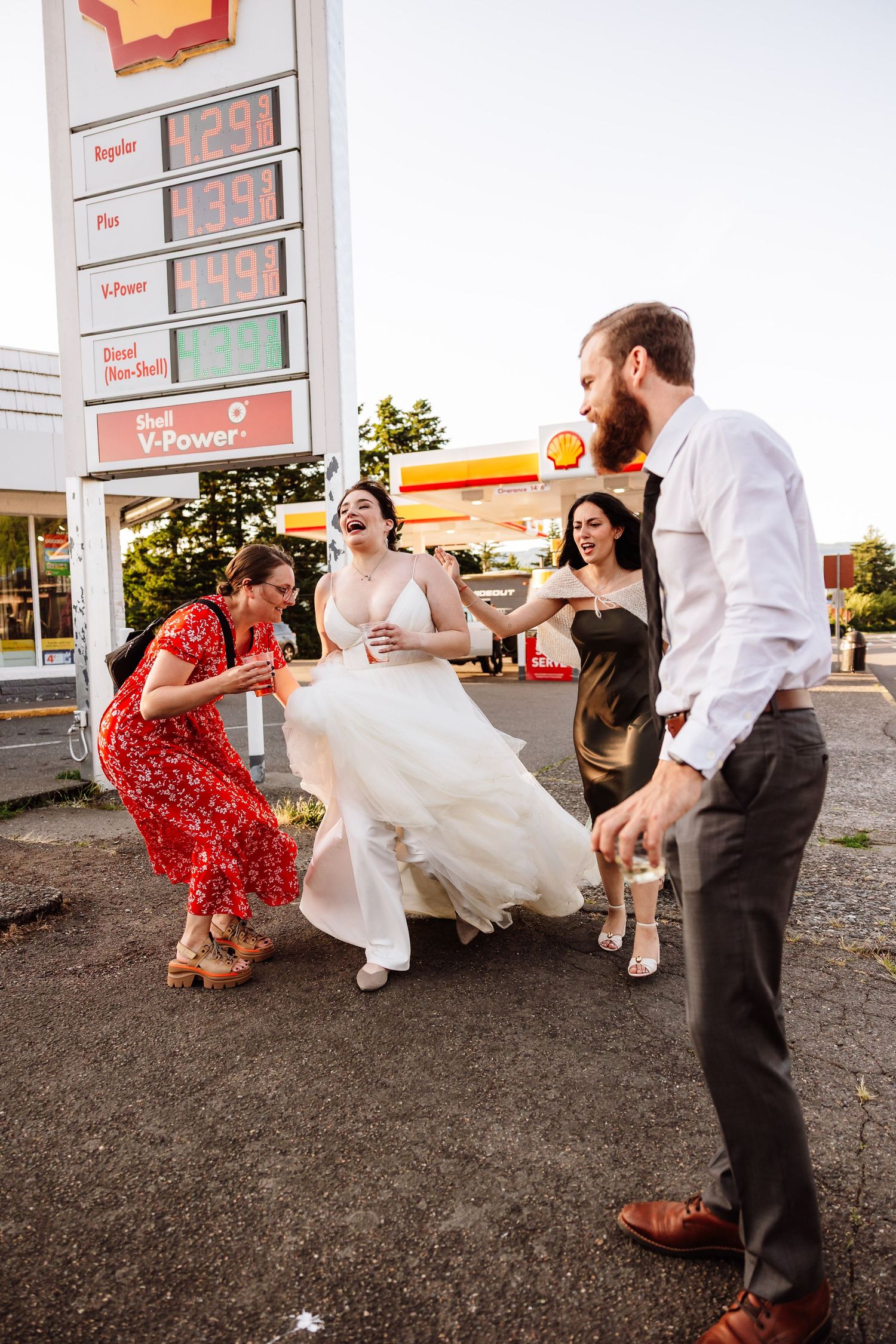 Bride laughing with wedding party outside a Shell gas station in a candid celebratory moment.