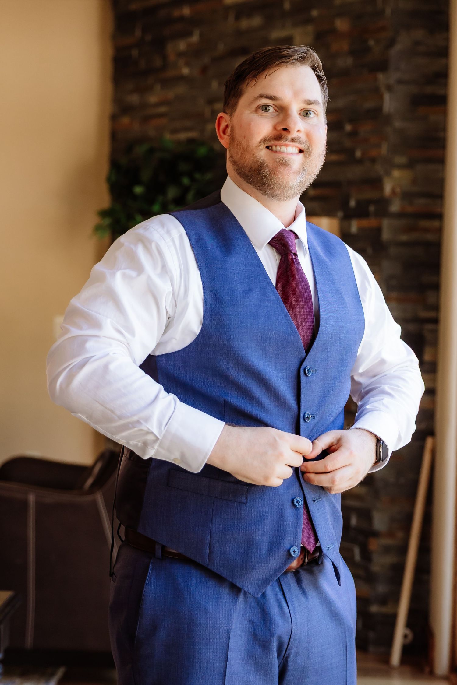 Smiling groom in blue vest and burgundy tie buttons his suit during wedding preparations.