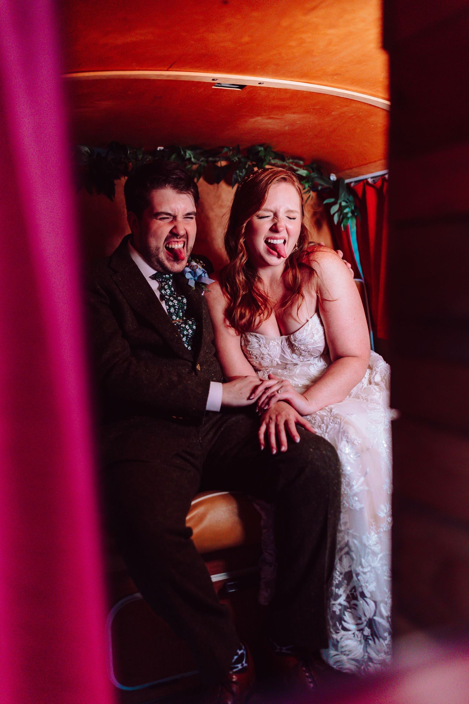 Bride and groom laugh and stick out tongues playfully while seated inside a vintage photo booth.